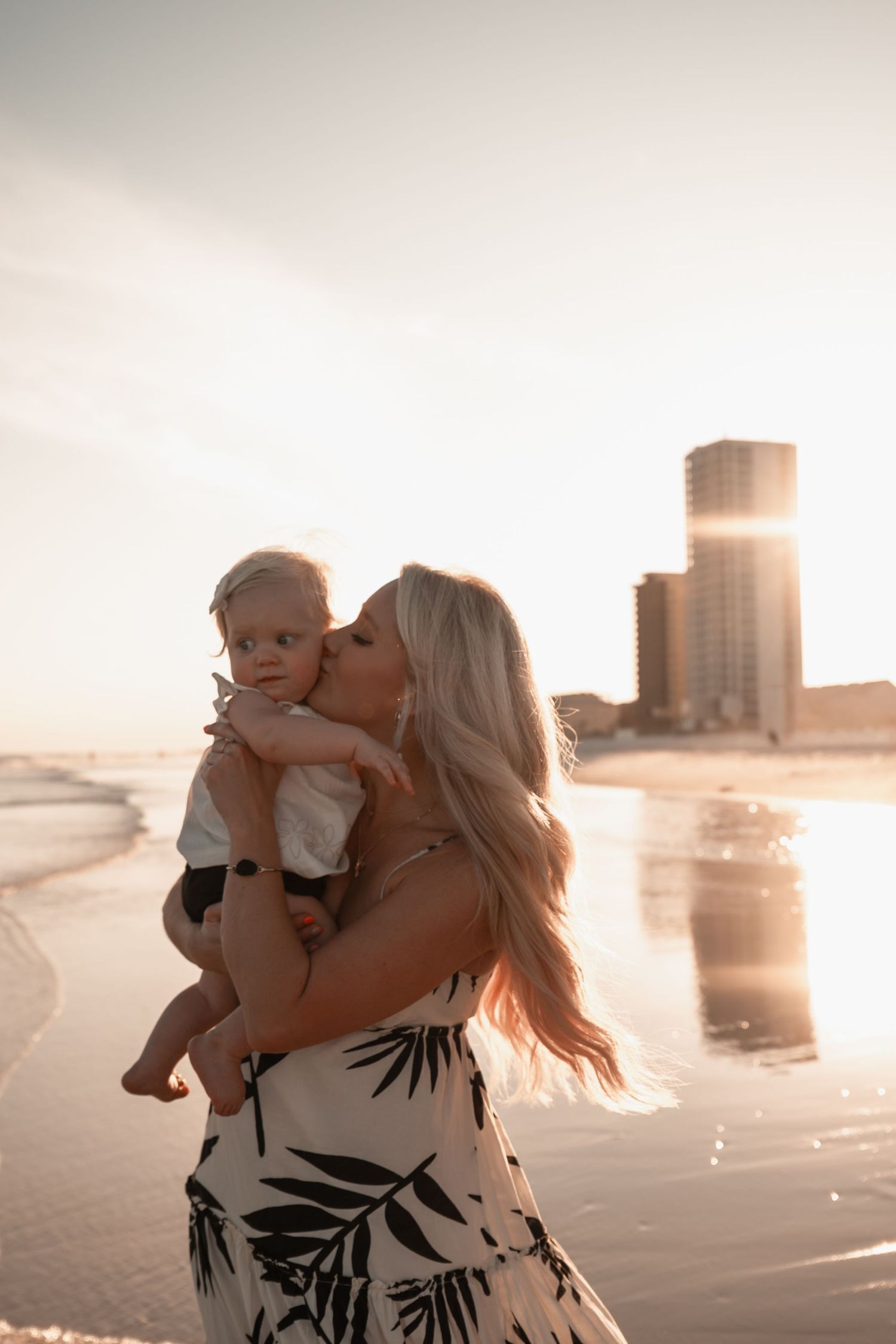 Blonde mother in floral dress kissing baby on a reflective beach at golden hour sunset.
