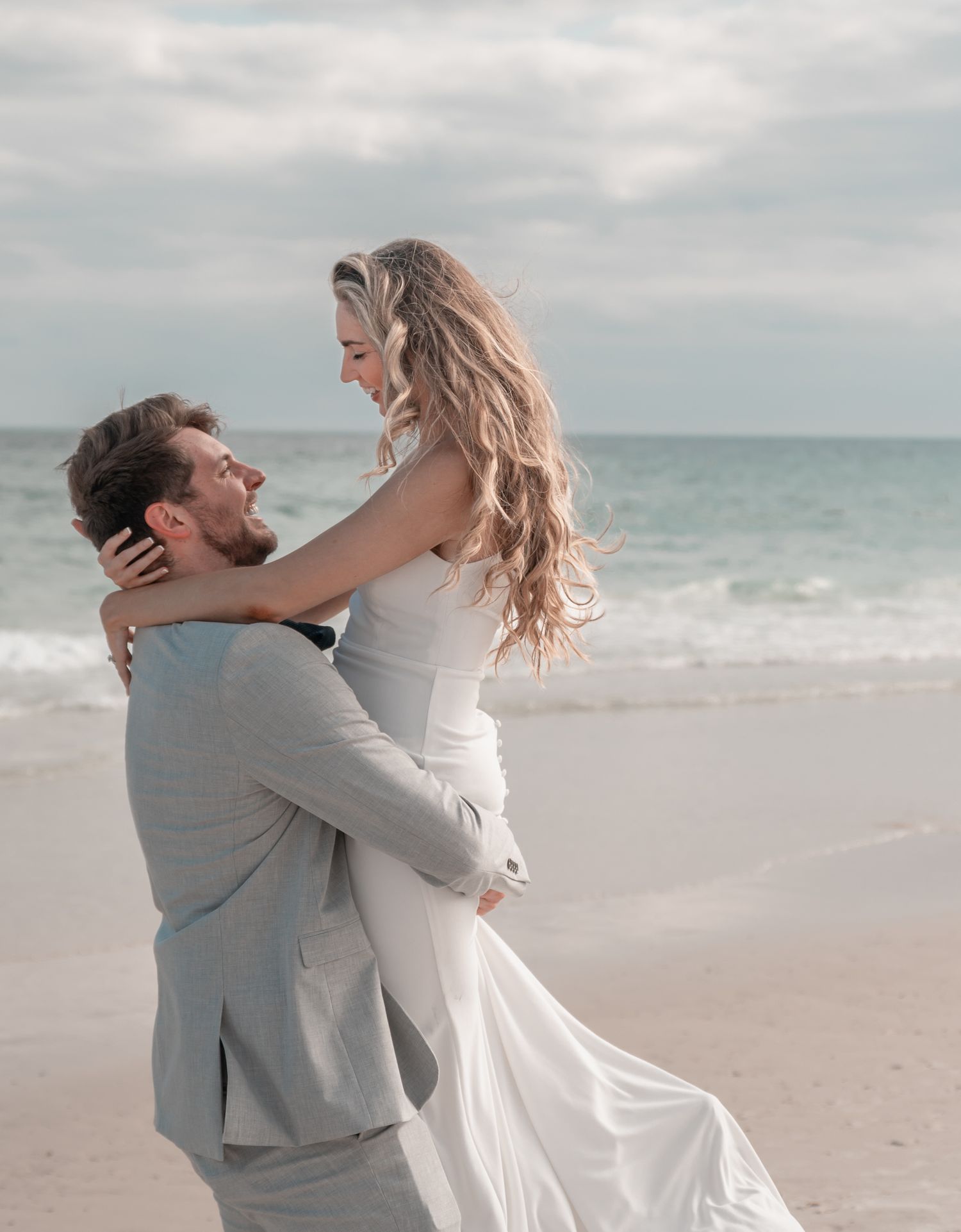 Groom in gray suit lifting bride in white wedding gown on a beach with ocean waves in the background.