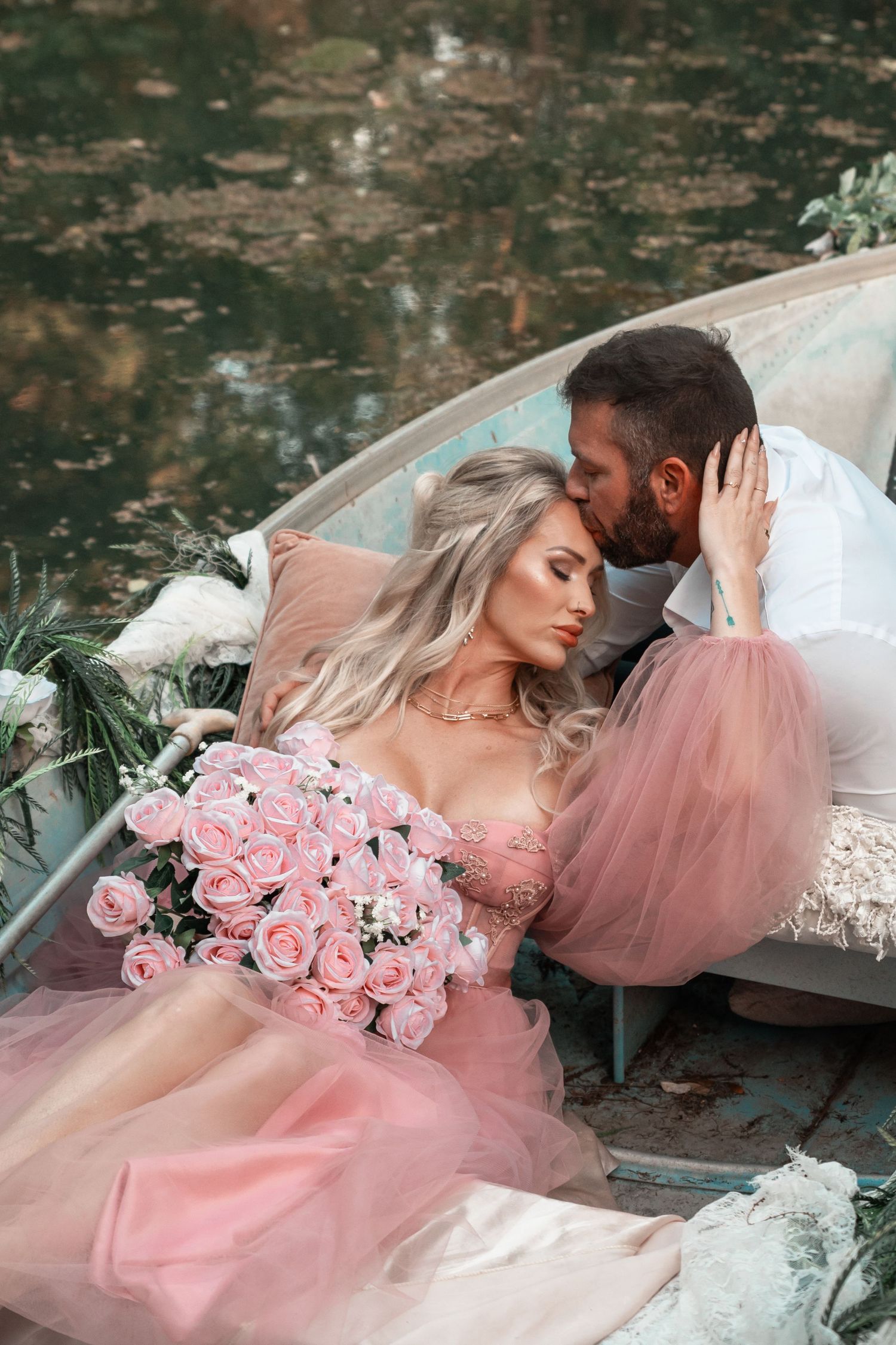 Blonde woman in pink tulle gown with bouquet kissed by bearded man on boat.