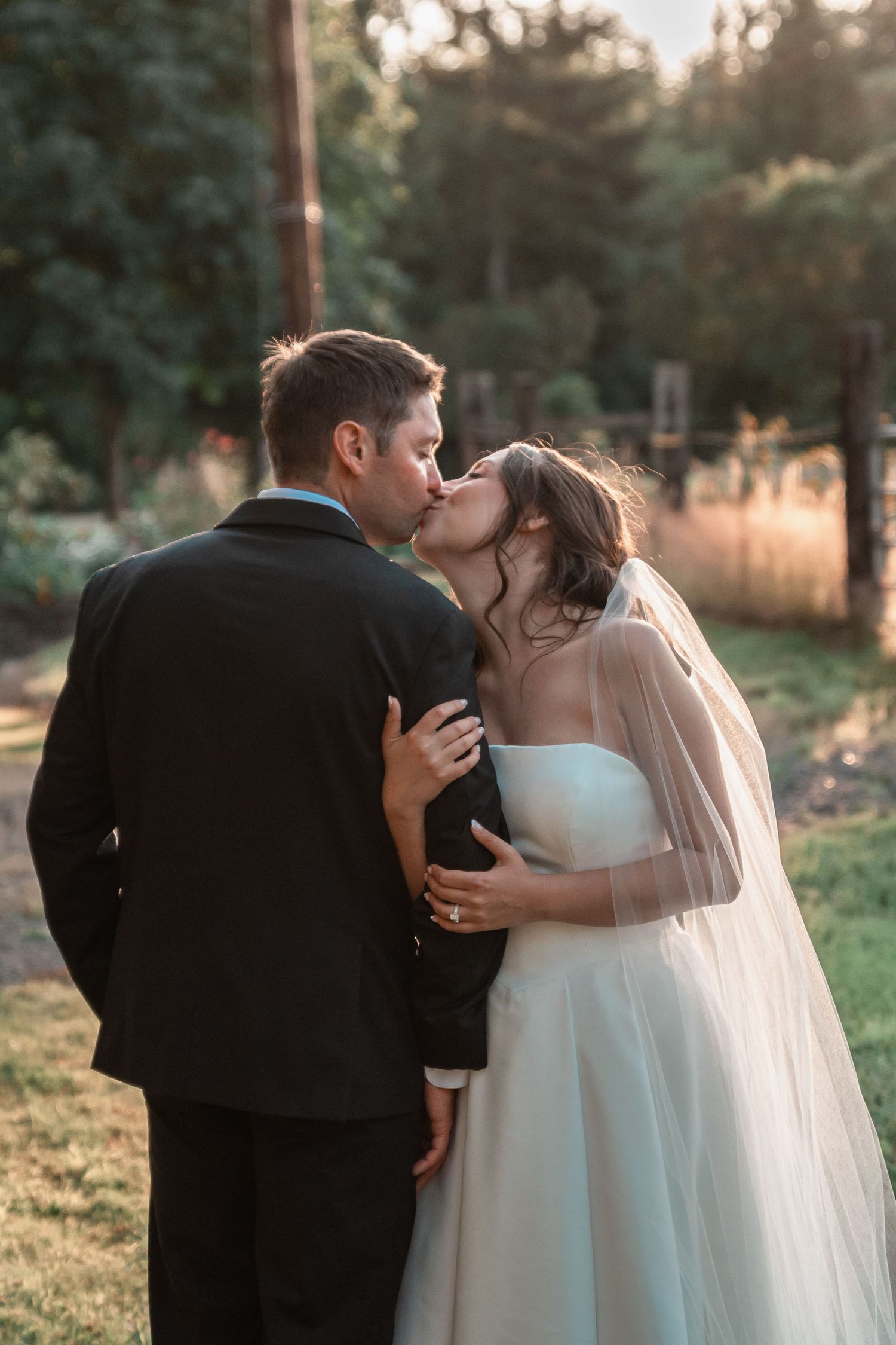 Bride and groom sharing a romantic kiss outdoors in golden hour light, bride wearing white gown with veil.