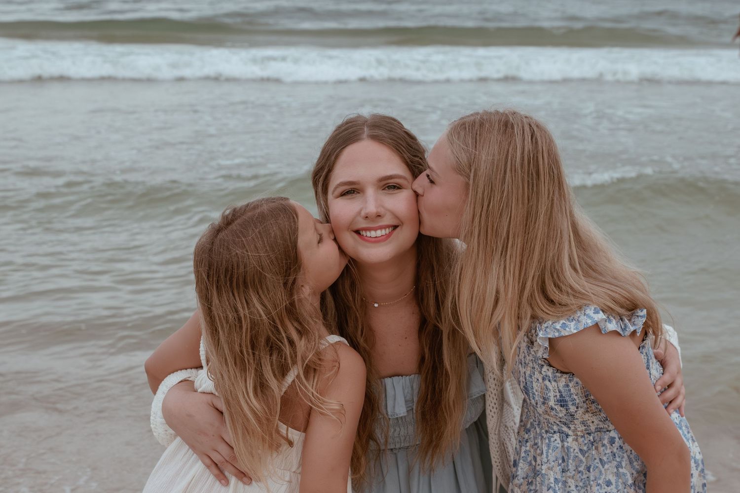 Three girls smiling on a beach, two younger girls kissing the cheeks of a teenage girl in a gray dress.