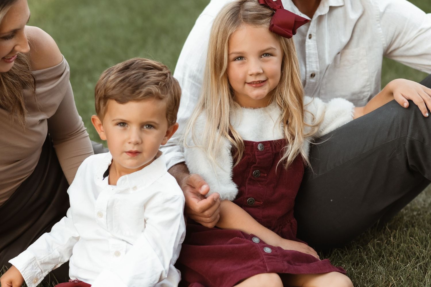 Two young siblings in burgundy and white outfits sitting together on green grass outdoors.