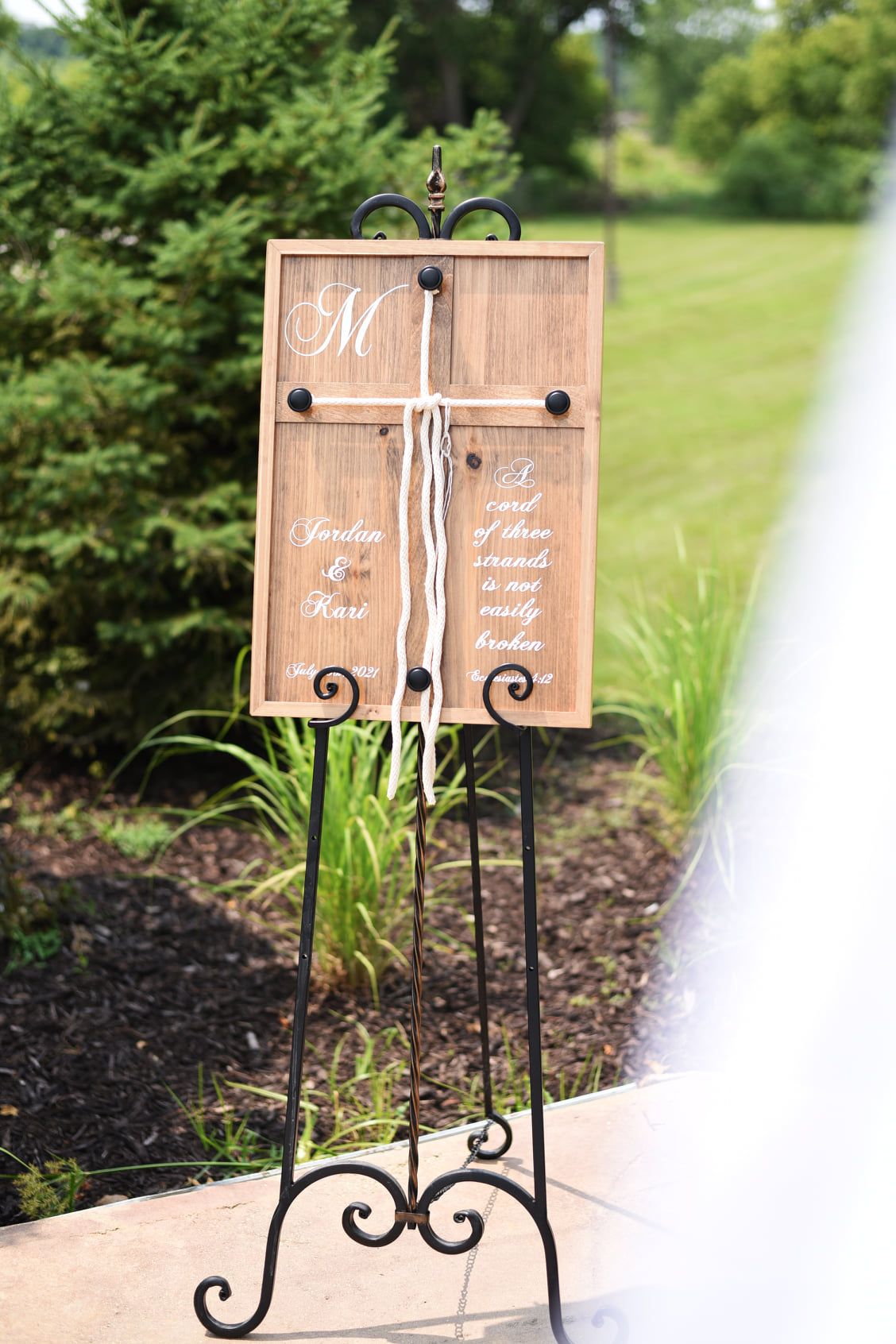 Rustic wooden wedding welcome sign on ornate black easel displaying monogram M and Jordan and Karl names outdoors.