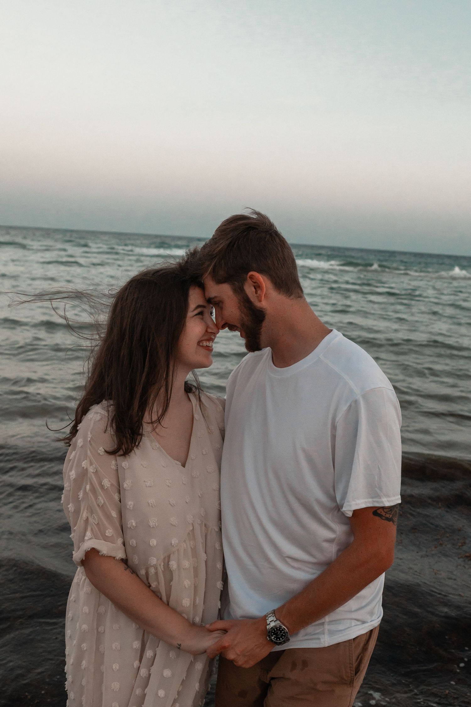 A romantic couple holds hands and touches foreheads on a beach at sunset, waves crashing behind them.