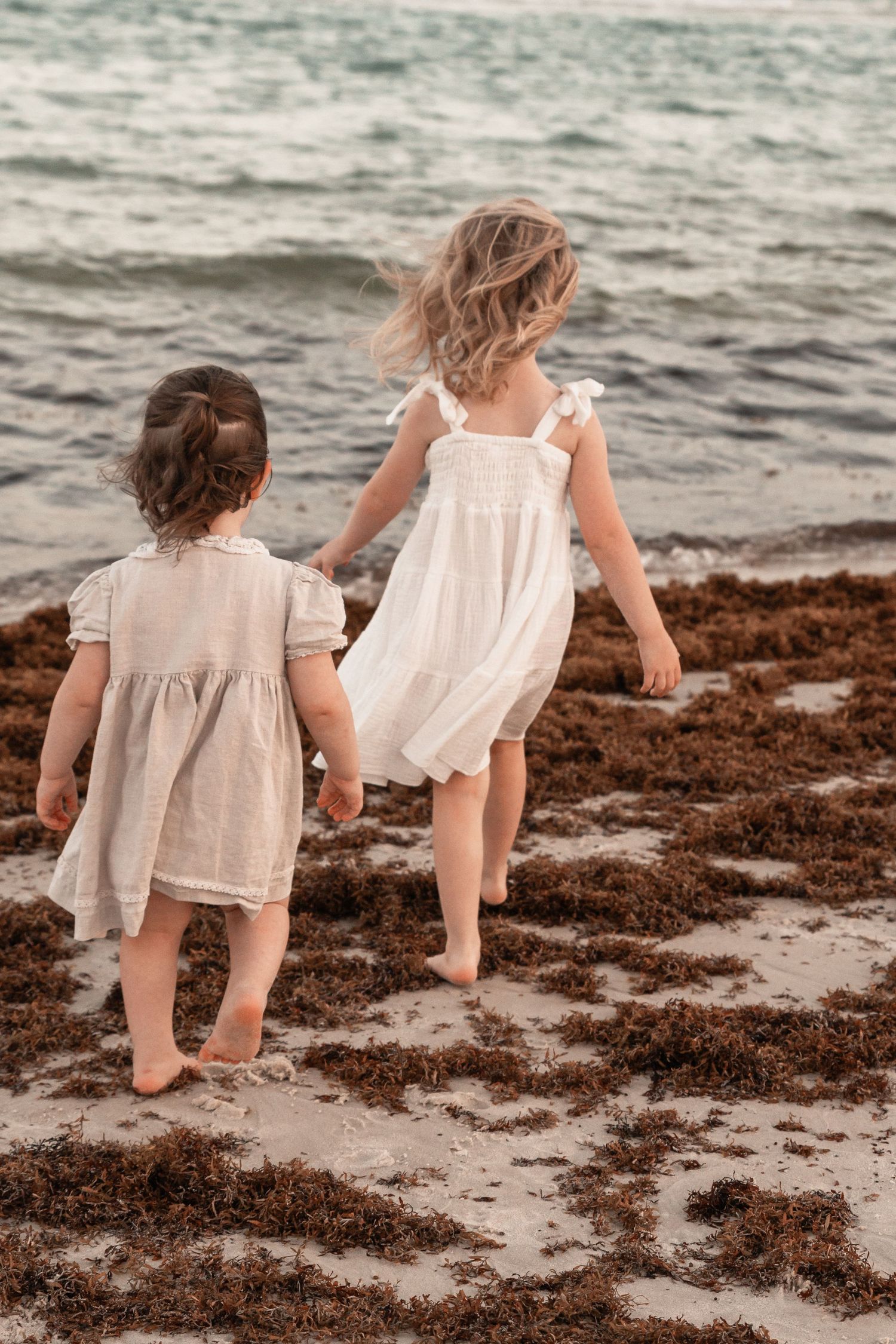 Two young girls in white dresses walk barefoot on a seaweed-covered beach toward the ocean waves.