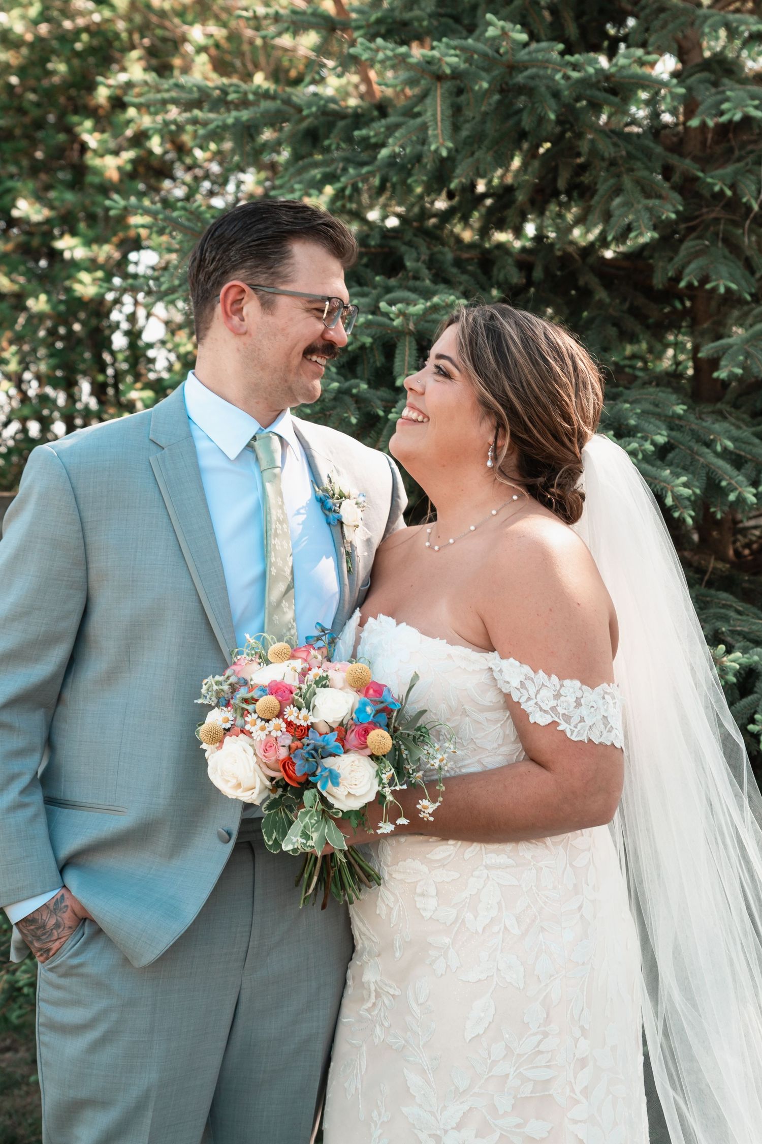 Smiling bride and groom in gray suit pose together outdoors near pine trees.