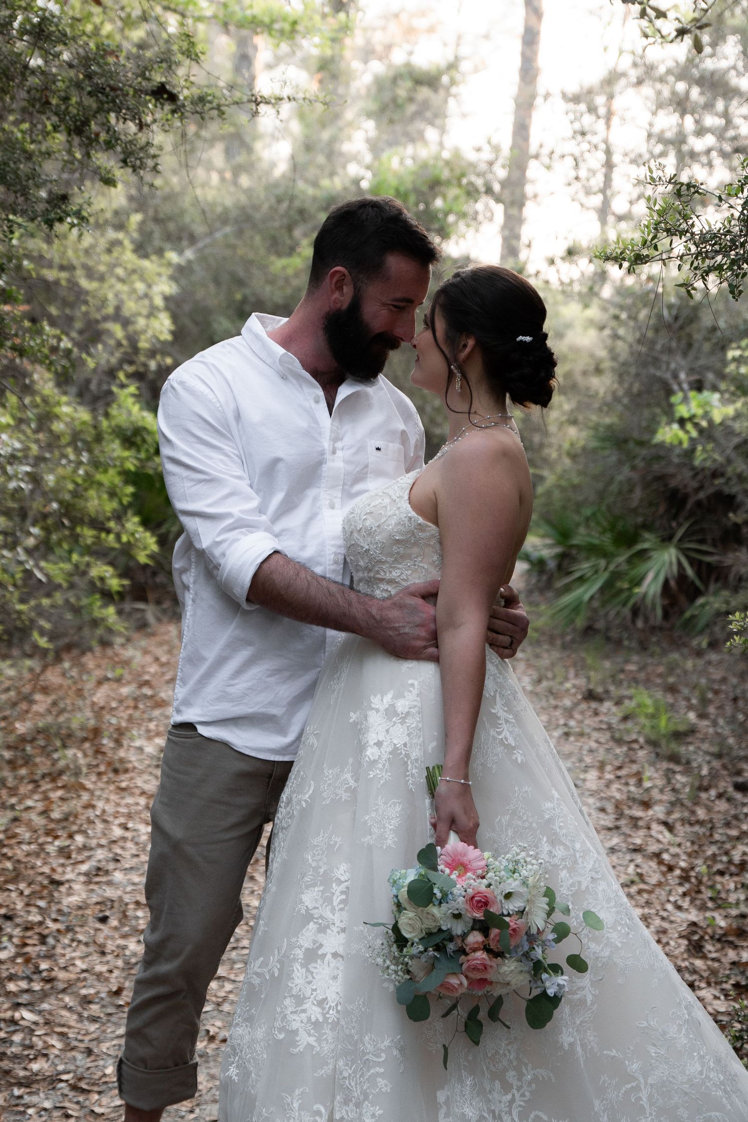Bride and groom share an intimate moment in a forest setting, she holds a pink and white bouquet in her lace gown.