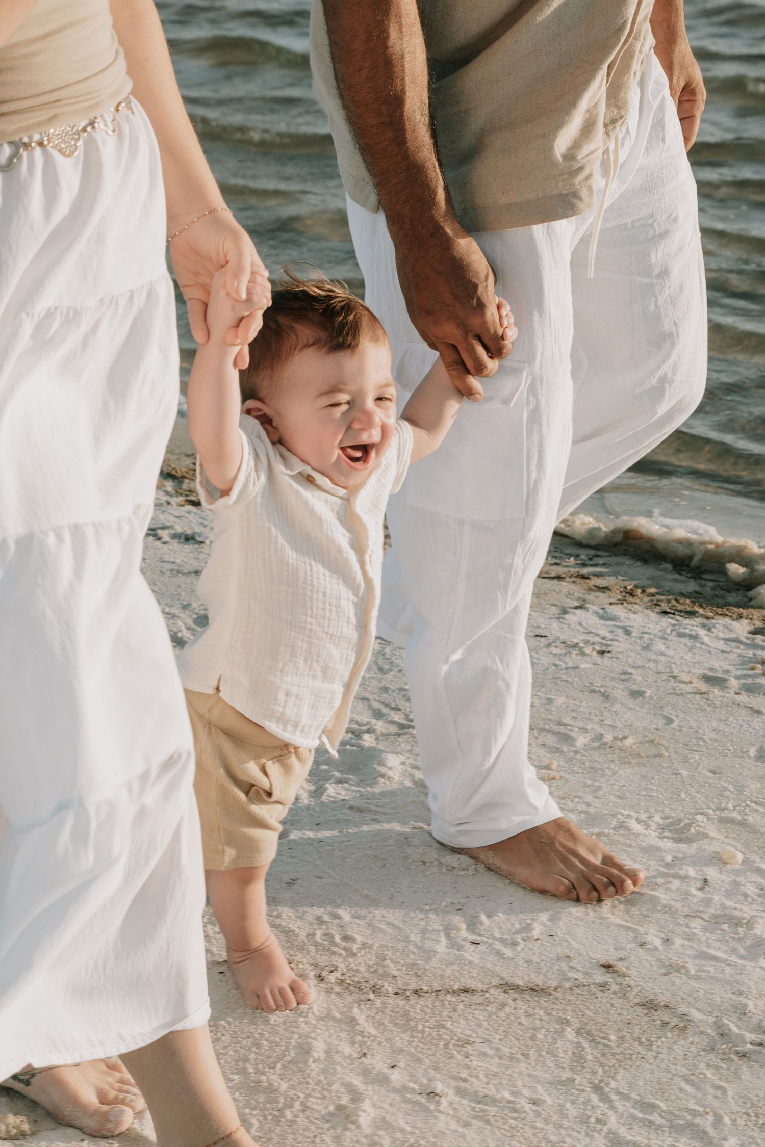 Laughing baby held by parents hands walking barefoot on beach shoreline.