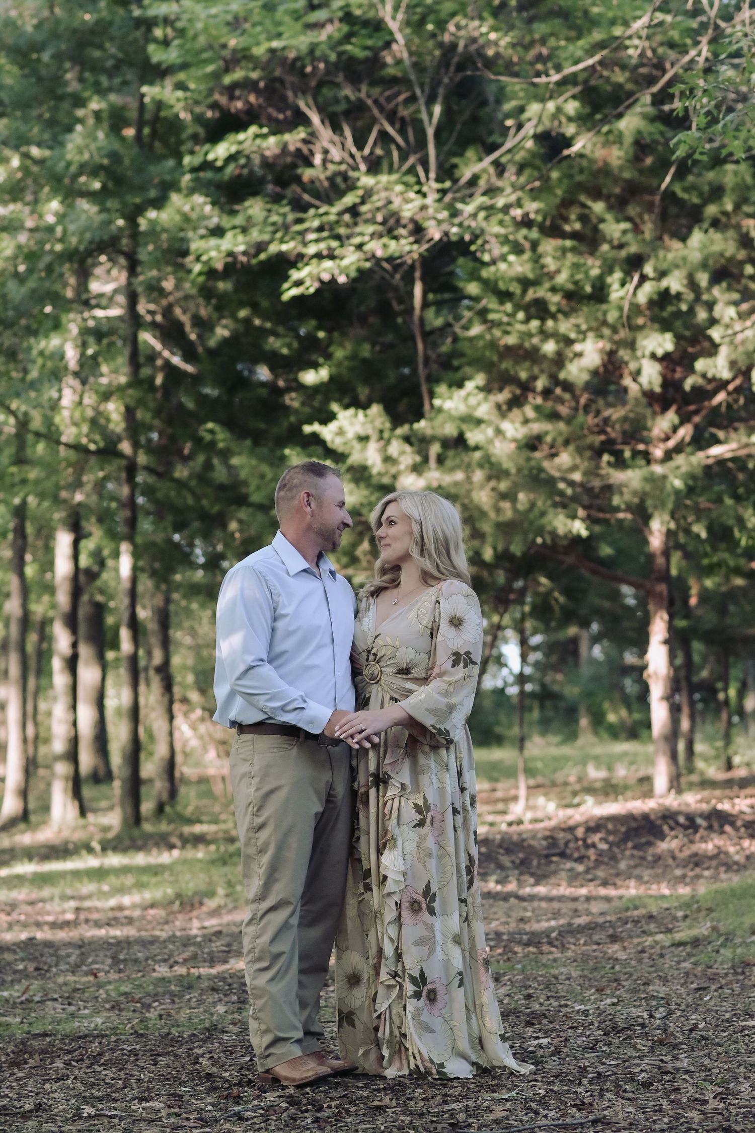 Engaged couple holding hands and gazing at each other in a sunlit pine forest during engagement session.