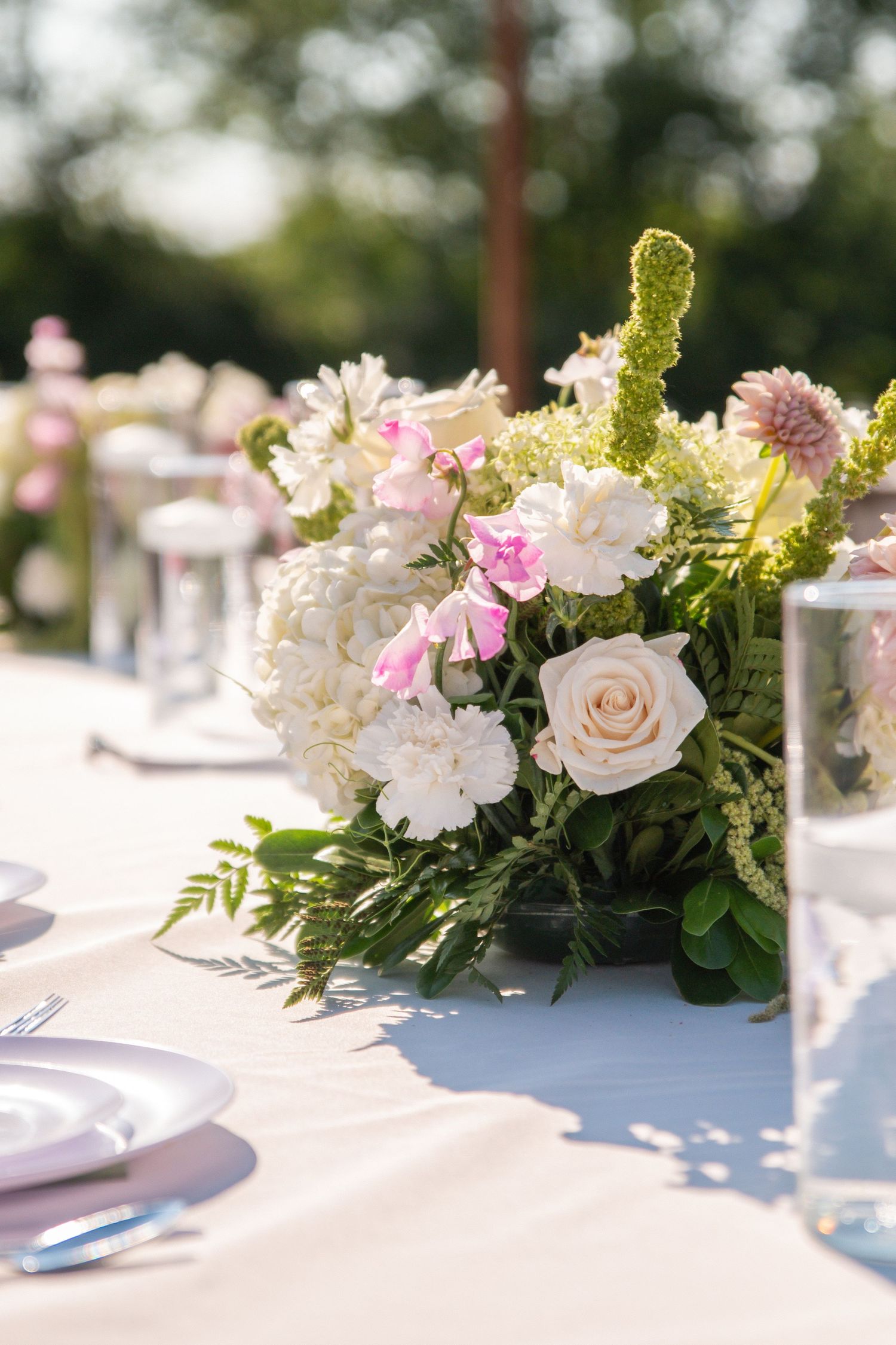Elegant outdoor wedding table with pink and white floral centerpiece featuring roses, hydrangeas, and greenery.