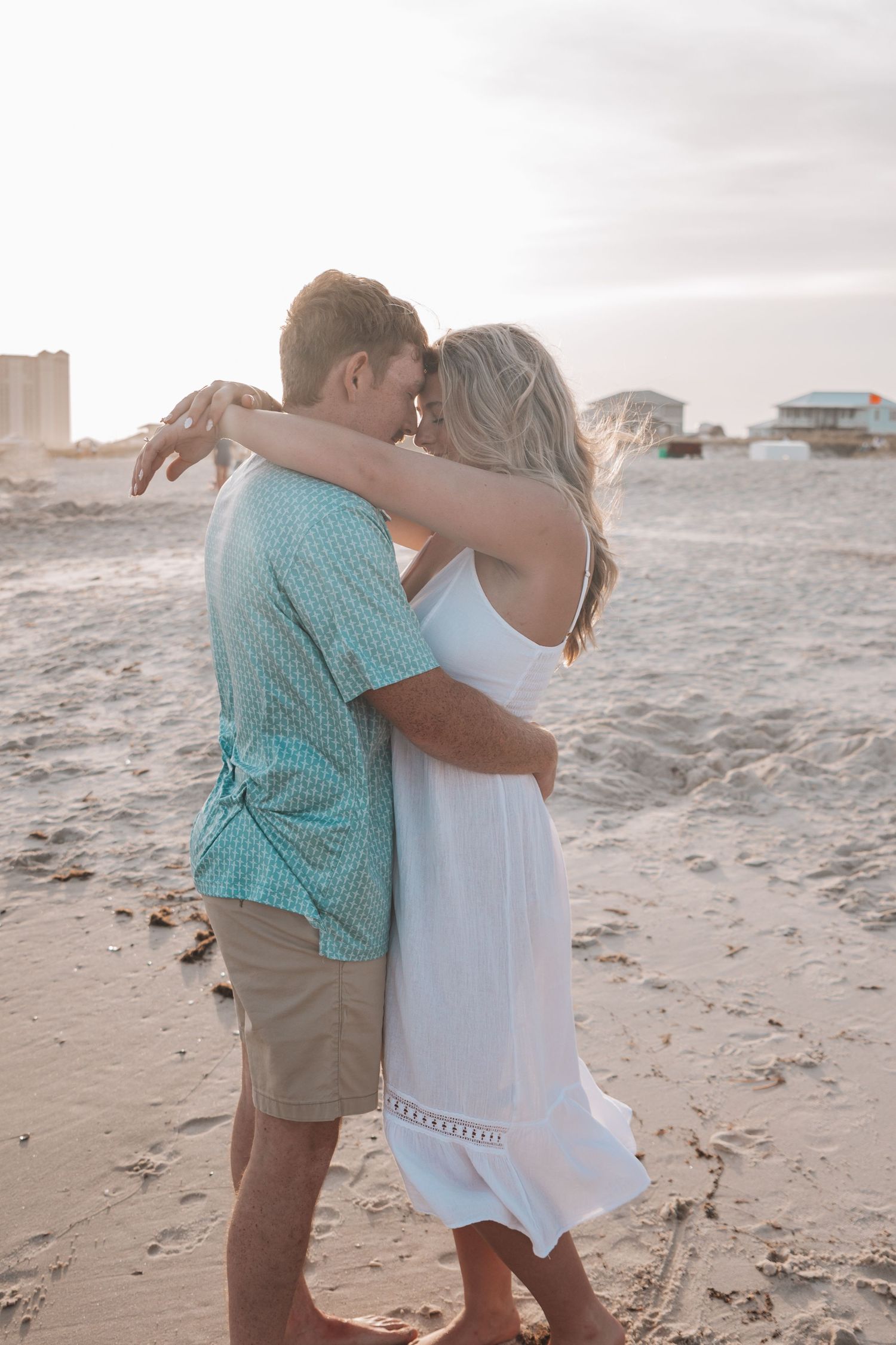Couple embracing romantically on a sandy beach at sunset, woman in white dress, man in teal shirt and khaki shorts.