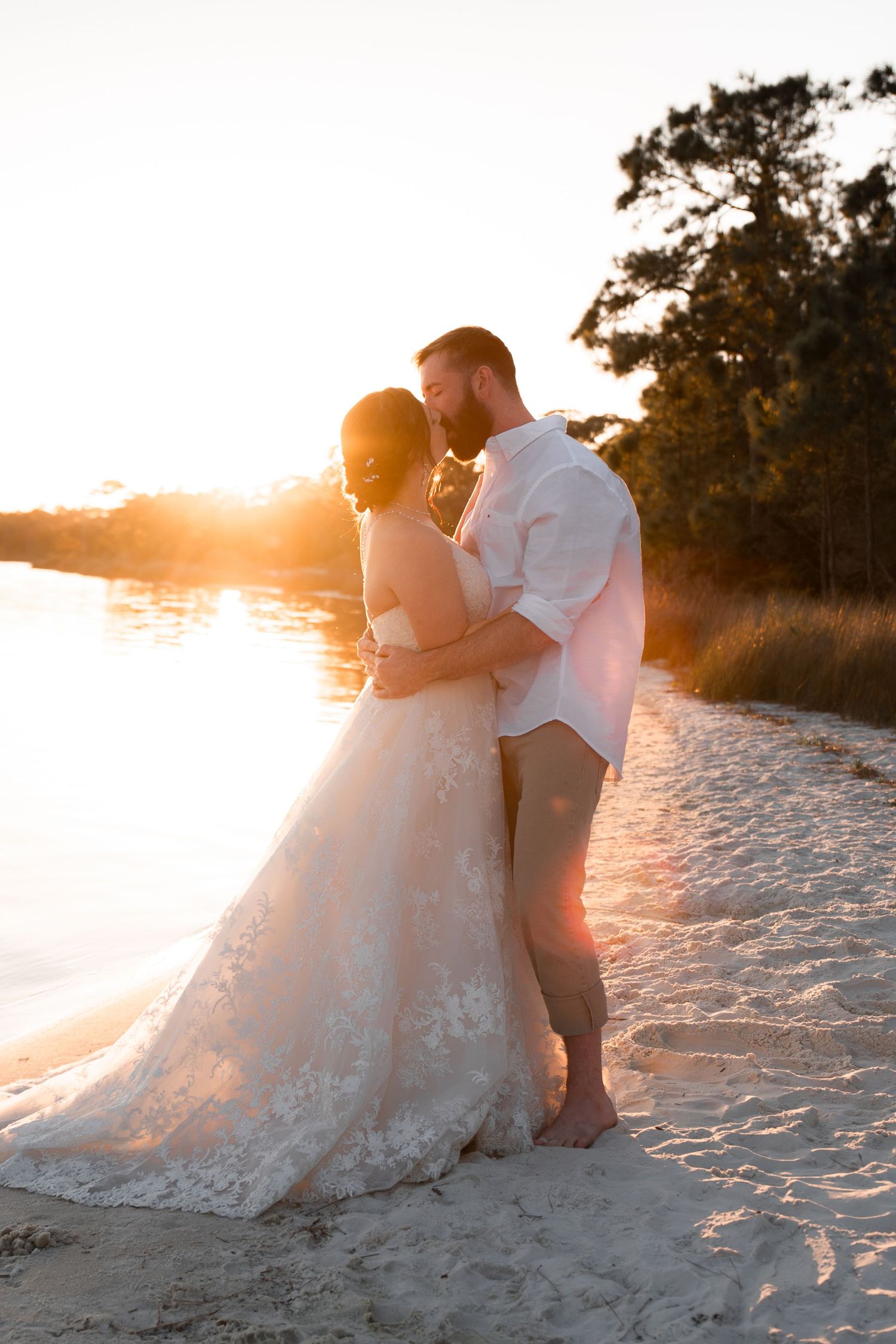 Bride and groom embracing on sandy beach at golden sunset, bride in lace gown, groom in white shirt barefoot.