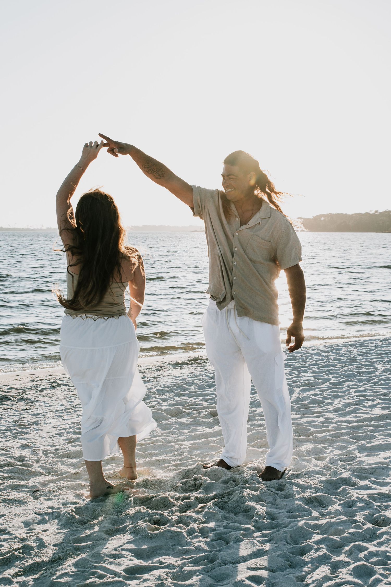 Couple dancing barefoot on sandy beach at golden hour, man spinning woman in white outfit.