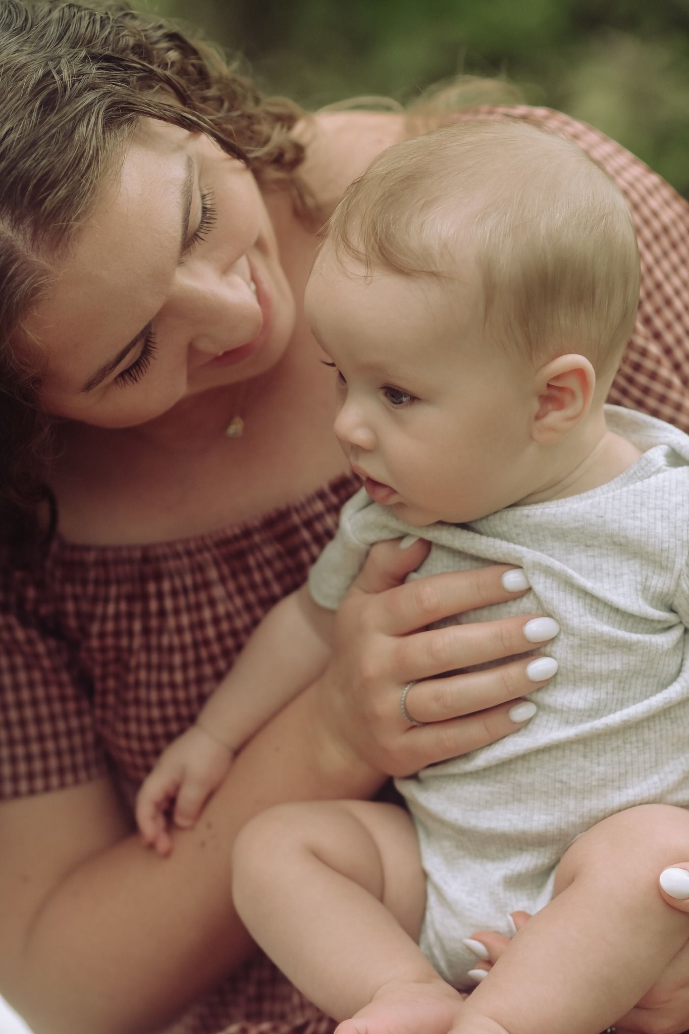 A curly-haired mother in a plaid dress smiles tenderly at her baby sitting in her lap.