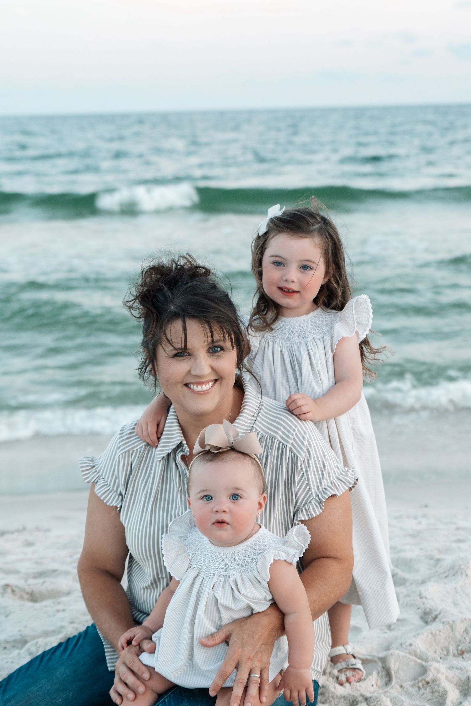 A smiling mother sits on a sandy beach holding a baby girl while a toddler in a white dress stands behind her.