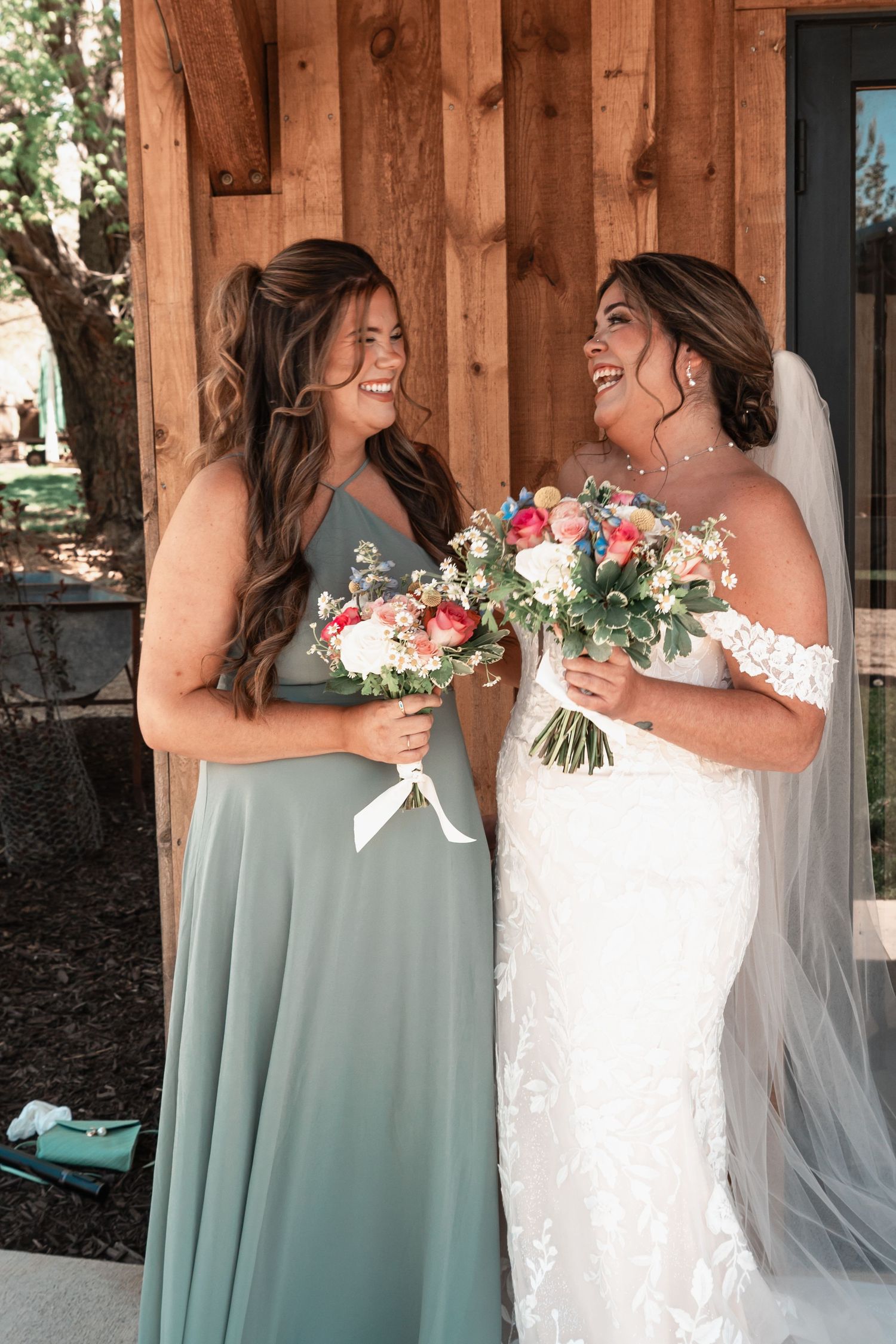 Bride in off-shoulder lace wedding gown smiles with bridesmaid in sage green dress outdoors.
