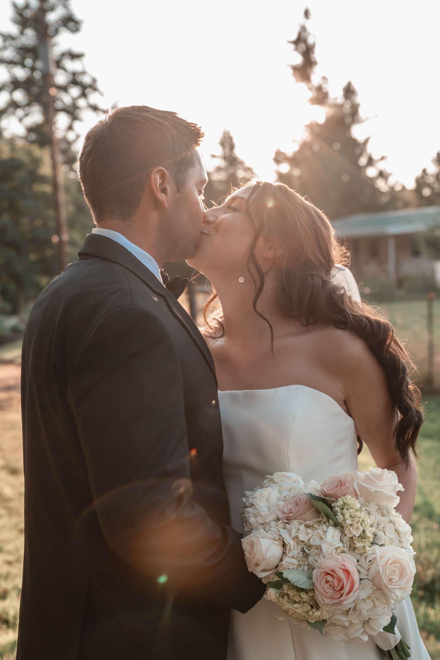Bride and groom sharing a romantic kiss at golden hour, bride holding a bouquet of blush pink roses and white flowers.
