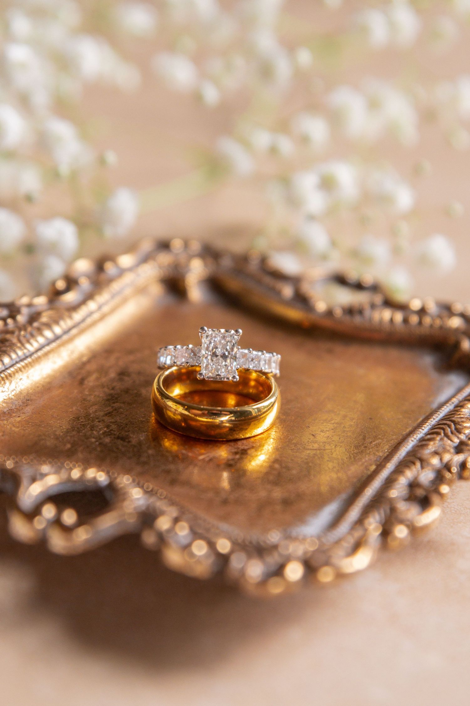 Gold wedding band and diamond engagement ring displayed on vintage ornate brass tray with white floral backdrop.