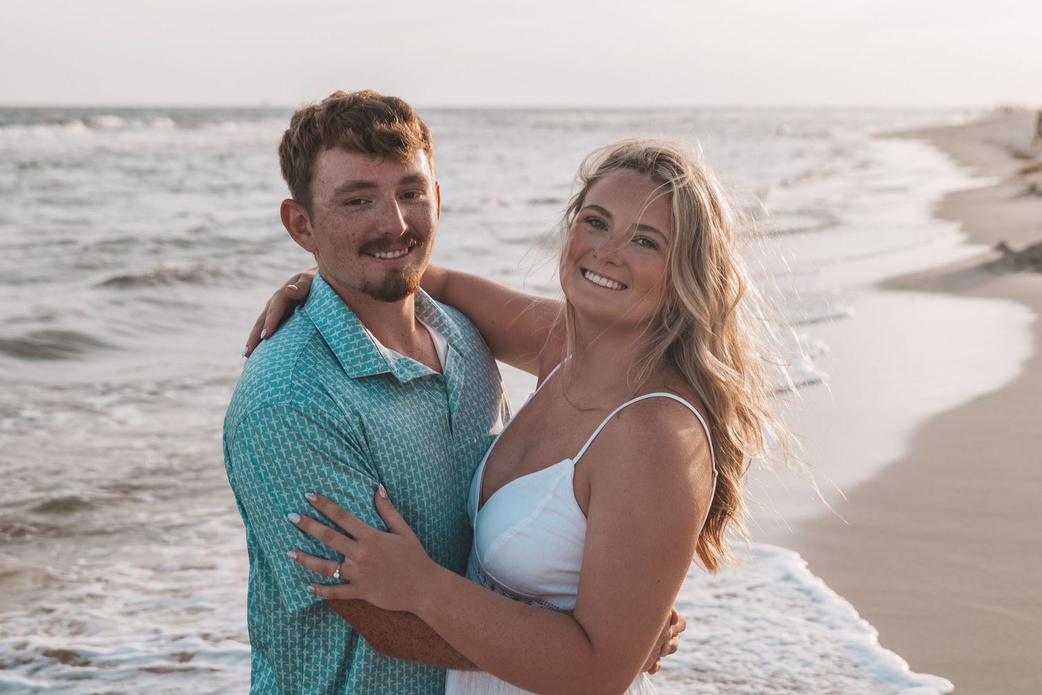 Happy couple embracing on a sandy beach at sunset, man in teal shirt and woman in white dress smiling.