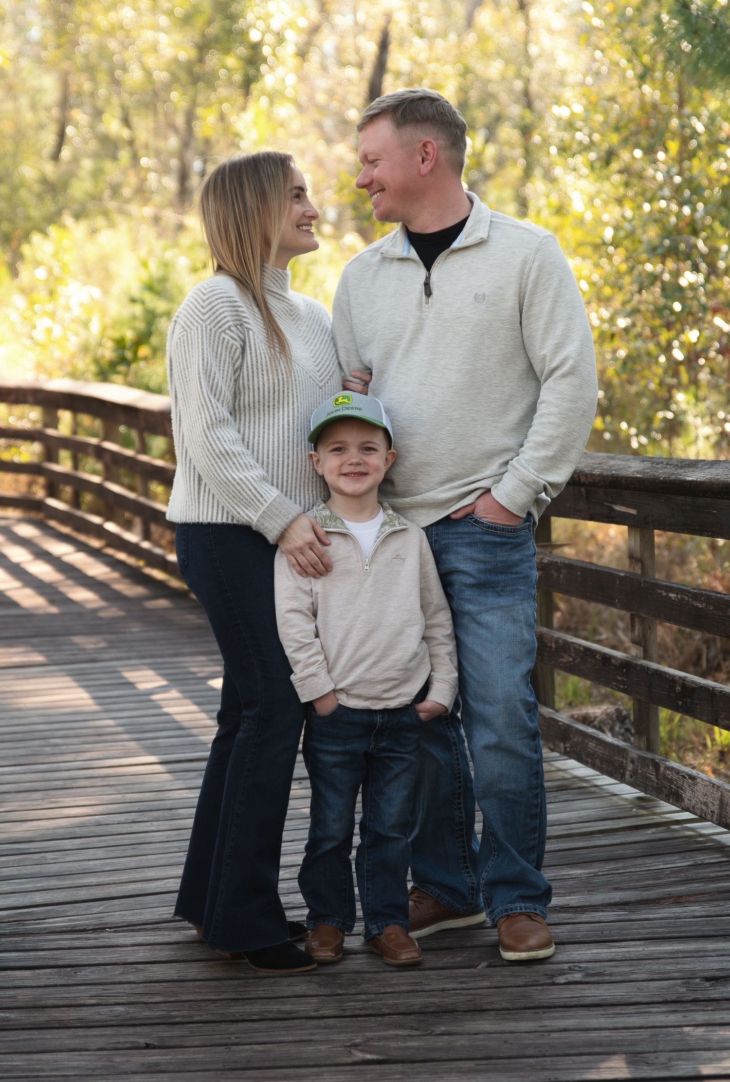 A smiling family of three poses on a wooden bridge surrounded by autumn foliage in a park.