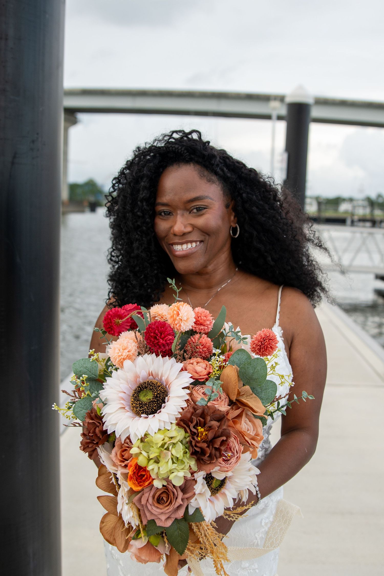 Smiling Black bride with curly hair holding a colorful fall bouquet with sunflowers and roses on a waterfront dock.