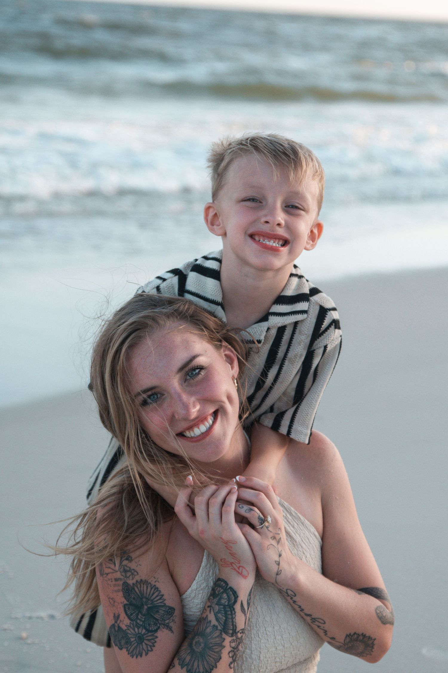 A smiling woman with tattoos carries a laughing young boy on her shoulders at a sandy beach with ocean waves.