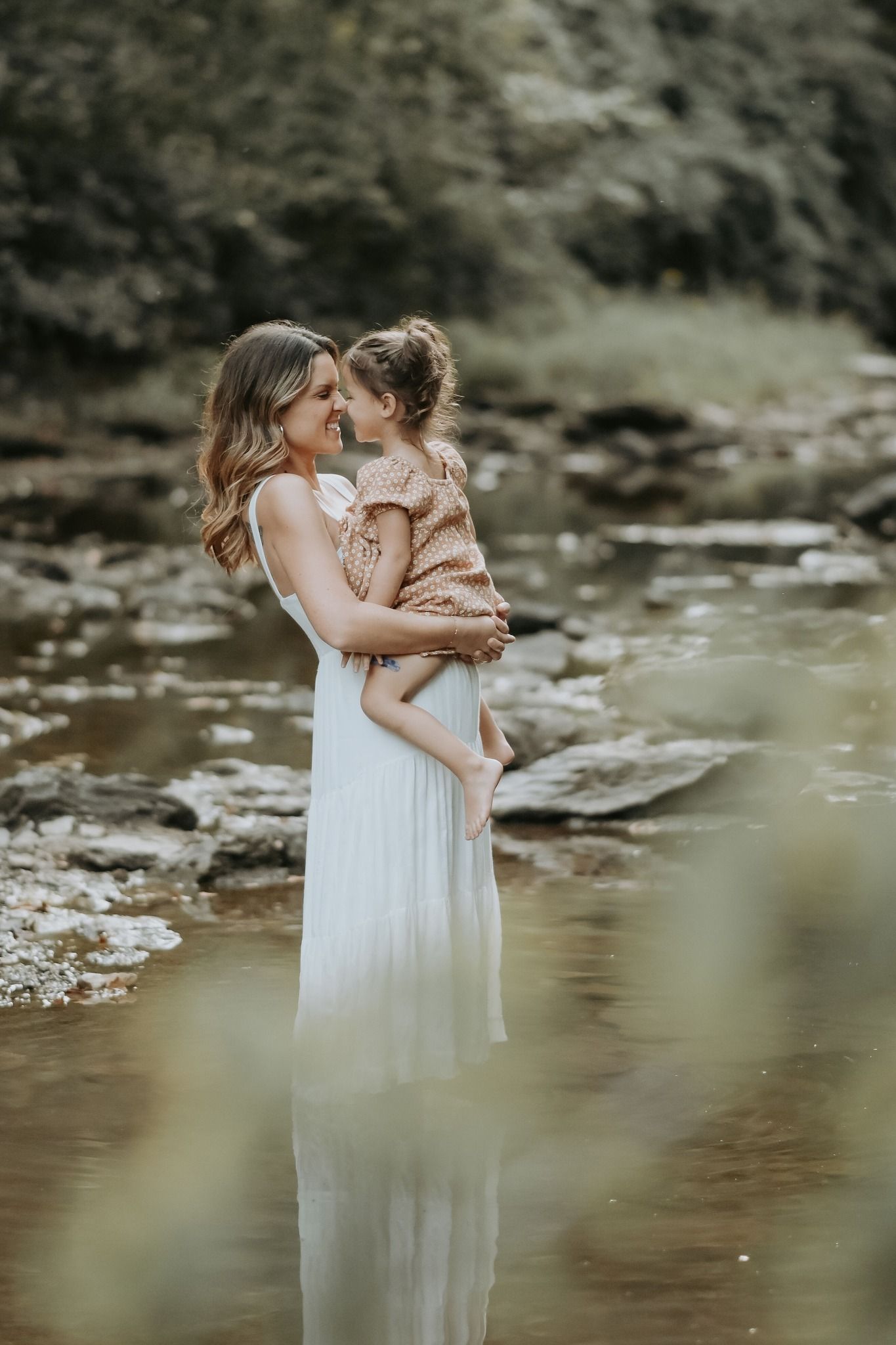 A mother in a white dress stands in a creek holding and smiling at her young daughter.