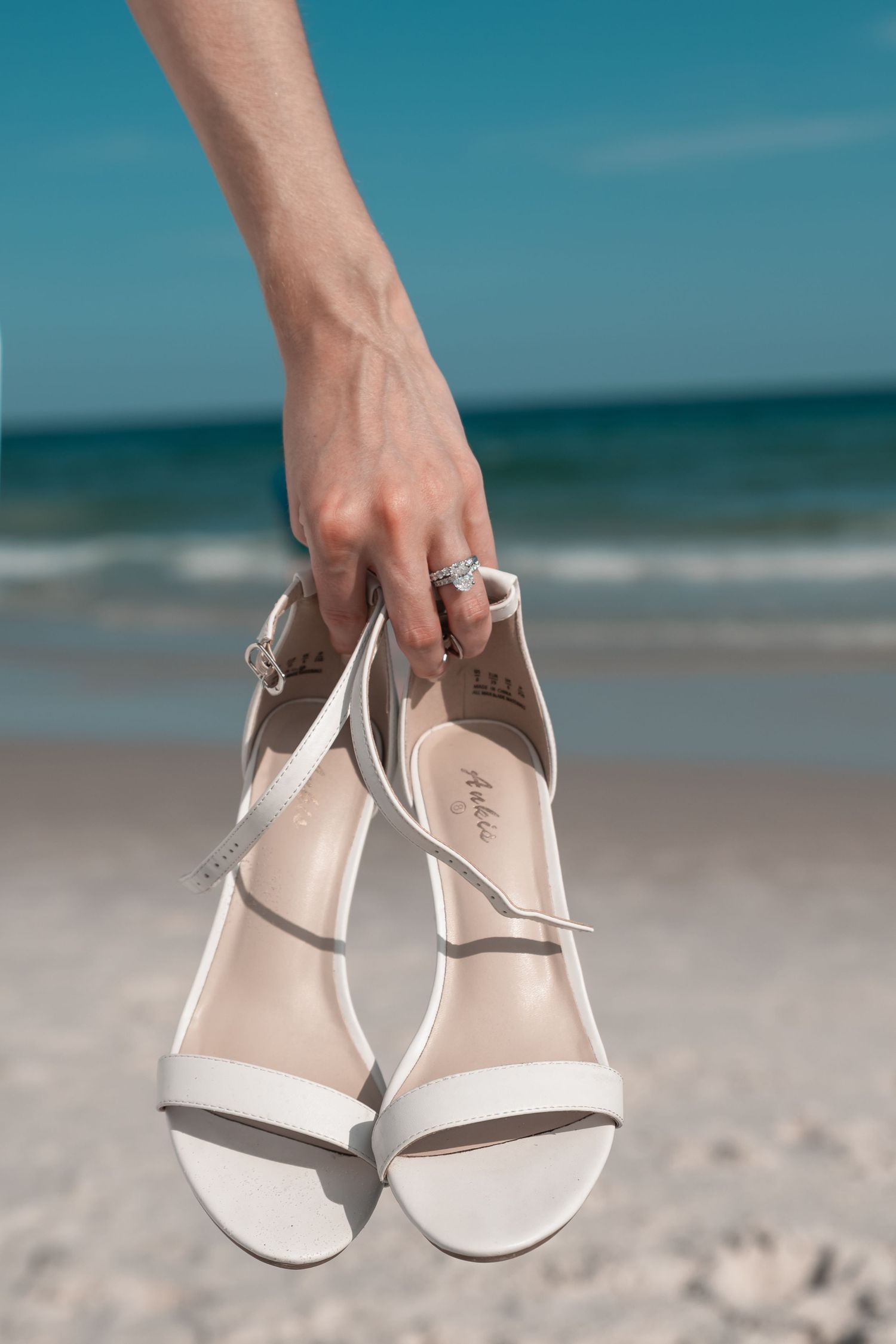 Hand holding white bridal sandals on a sunny beach with ocean waves in the background.