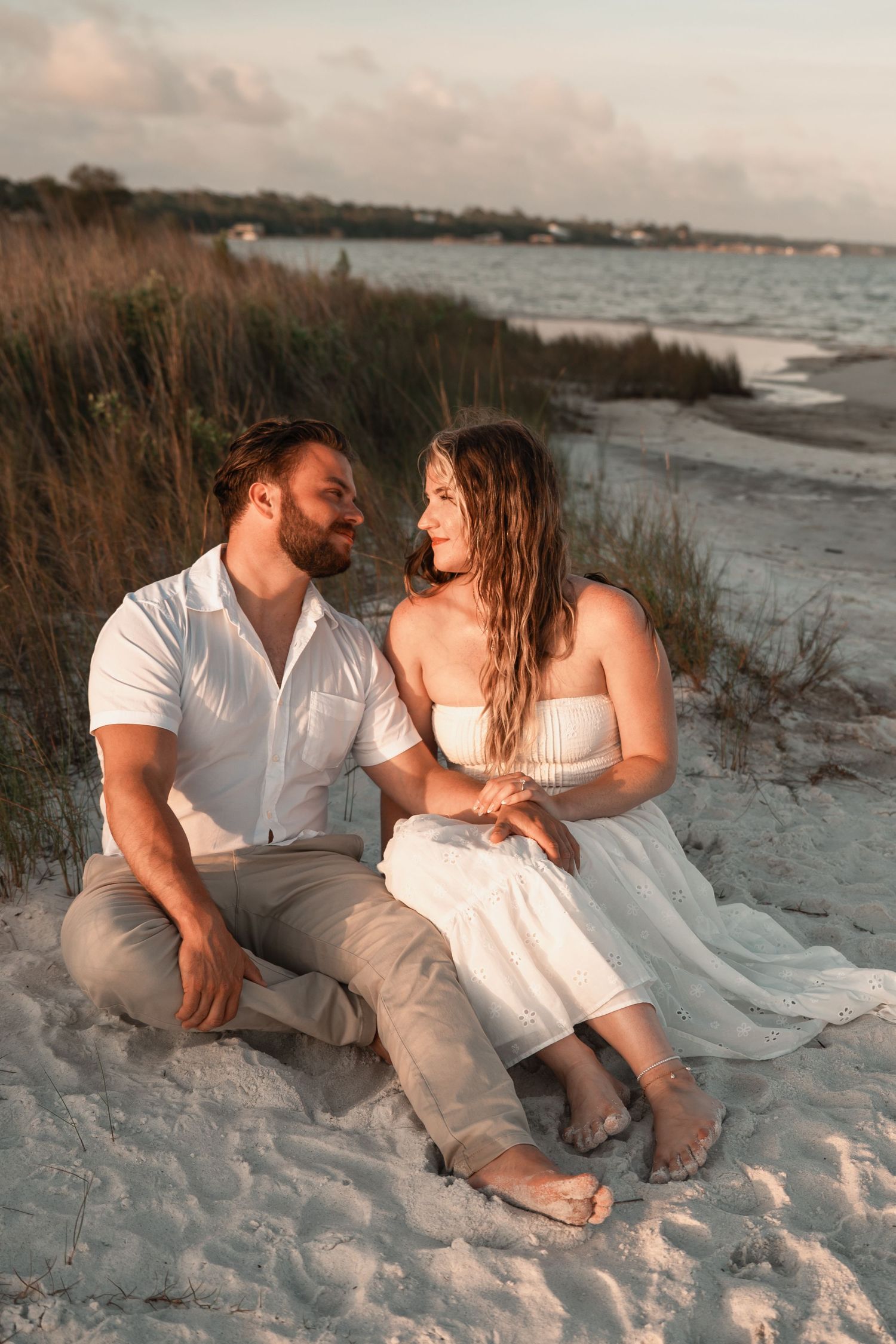 Couple sitting together on white sand beach at sunset, gazing into each others eyes near the water.