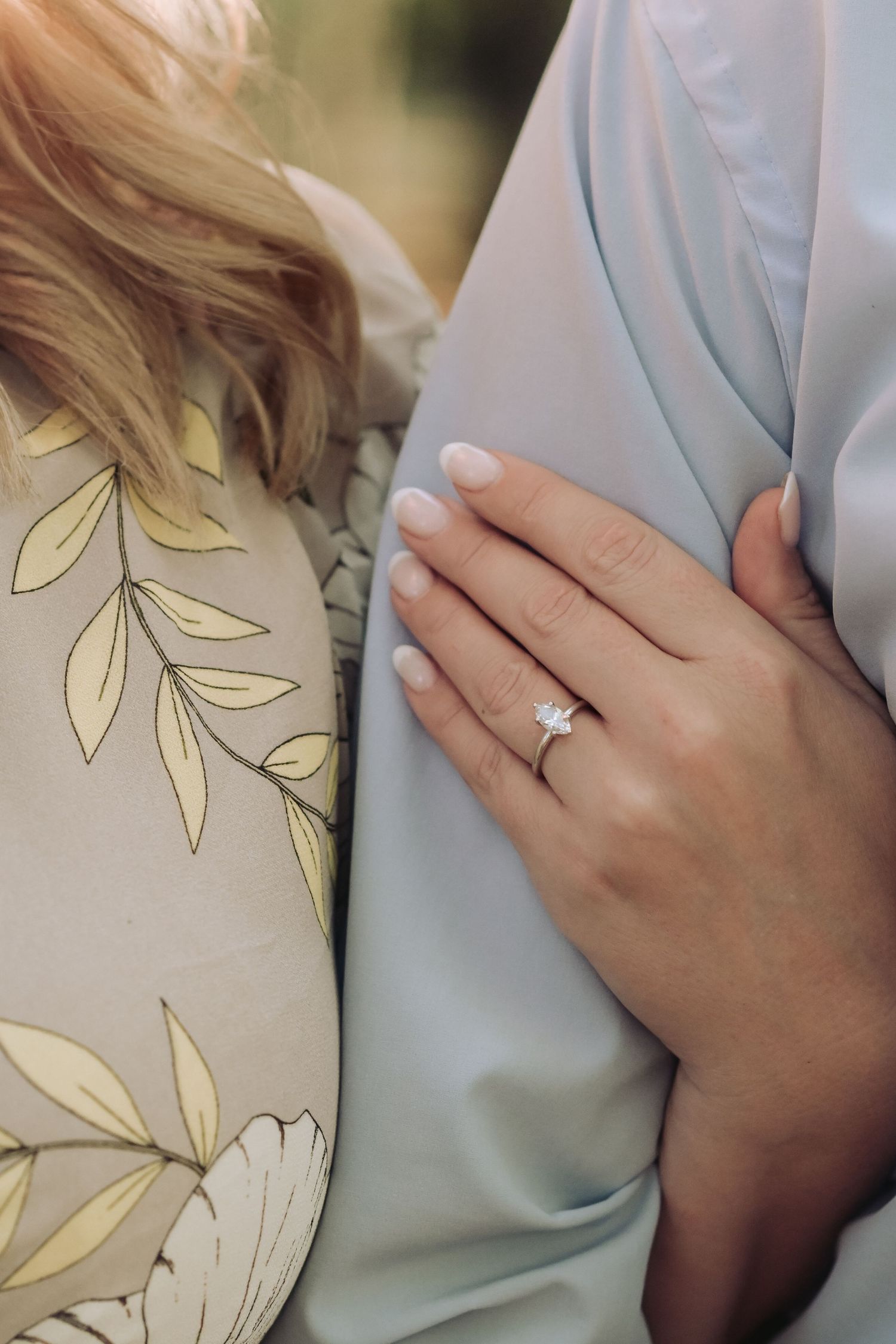 Close-up of a woman's hand displaying a solitaire diamond engagement ring while holding her partner's arm.