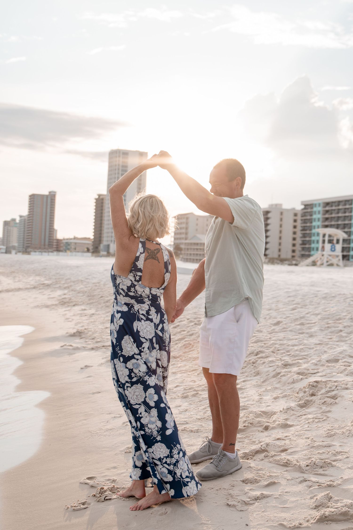 Couple dancing on a sandy beach at sunset with city skyline in the background, woman in floral maxi dress.
