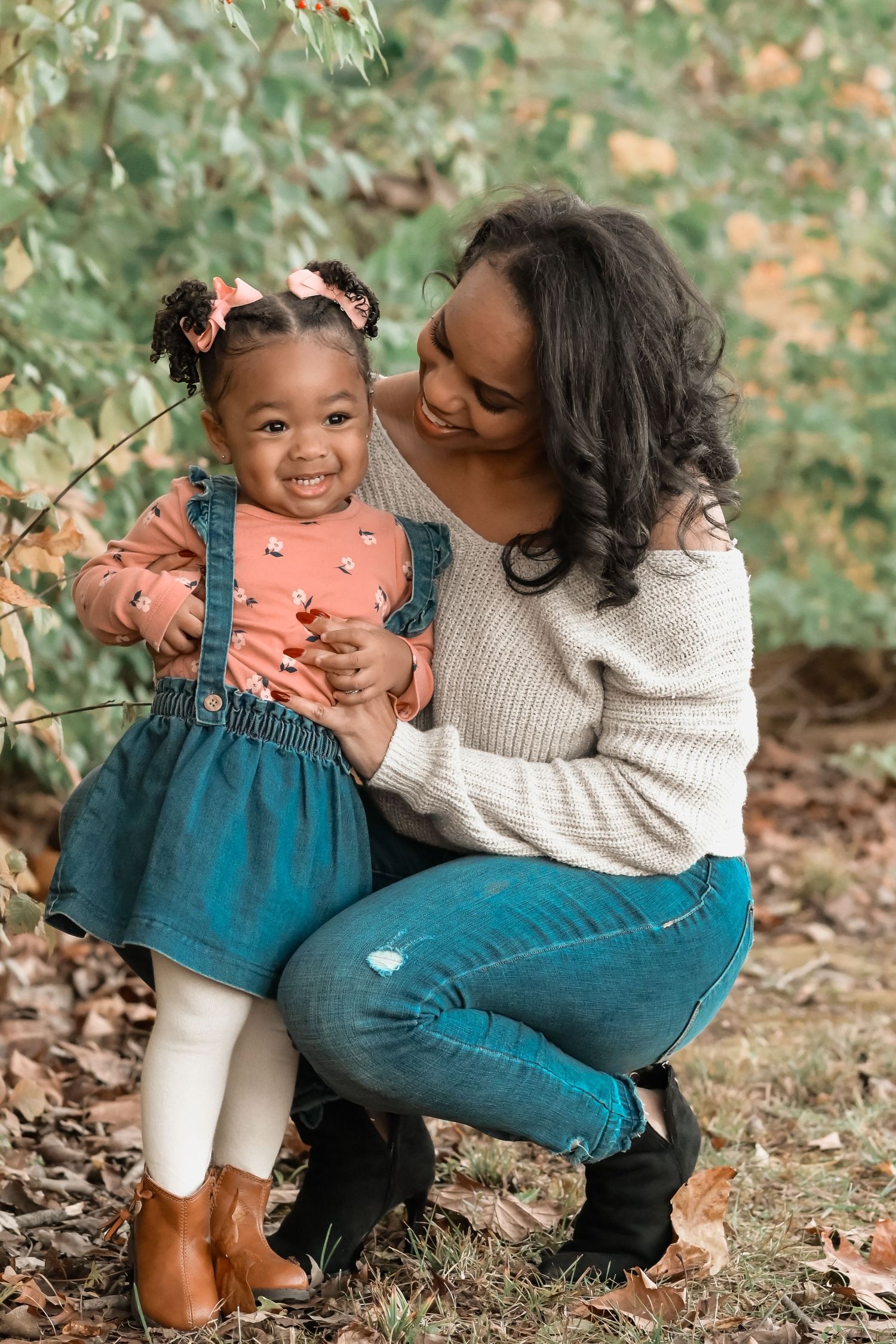 Black mother crouching with young daughter in autumn park, both smiling warmly.