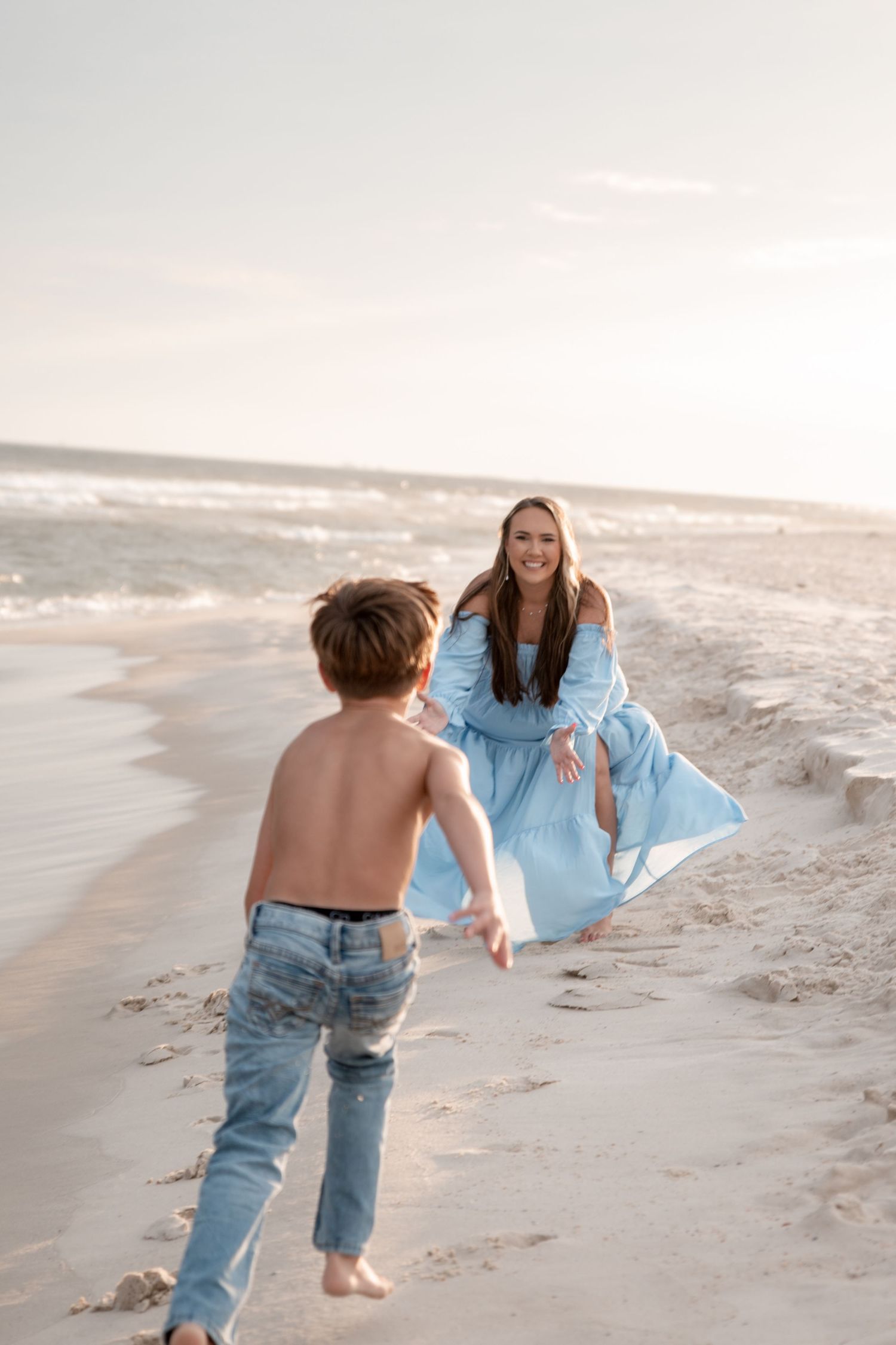 A smiling woman in a blue dress crouches on a sunny beach as a shirtless boy in jeans runs toward her.