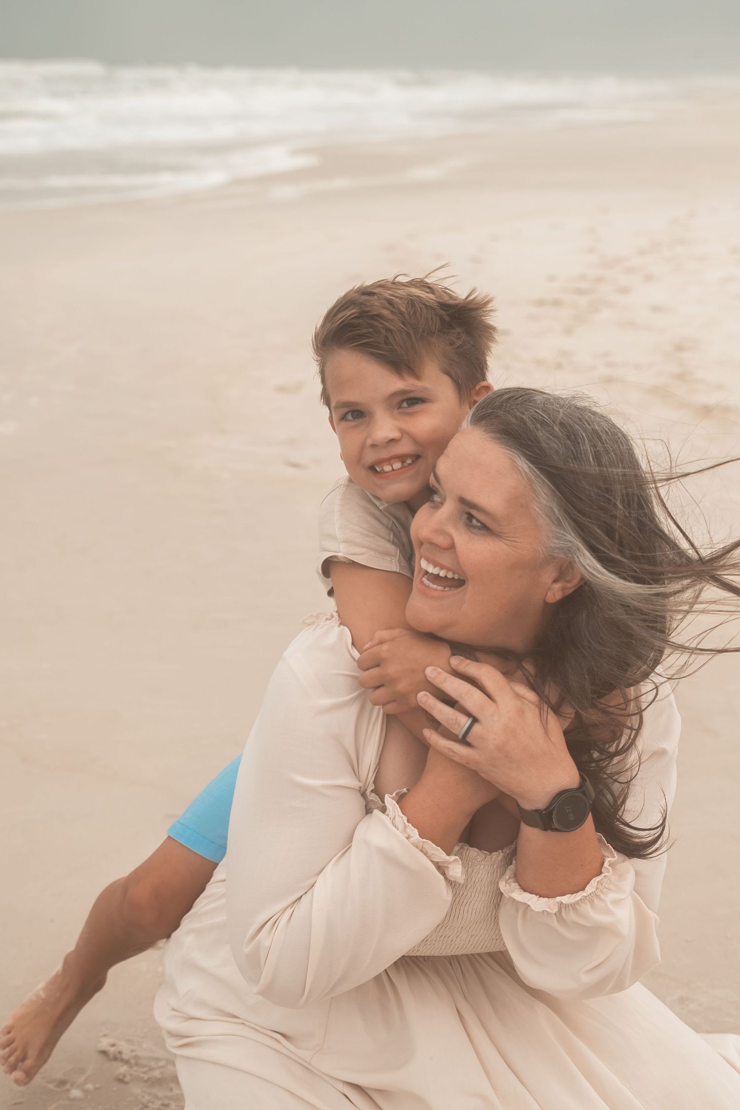 A grandmother and young boy share a joyful piggyback hug on a sandy beach with ocean waves in the background.