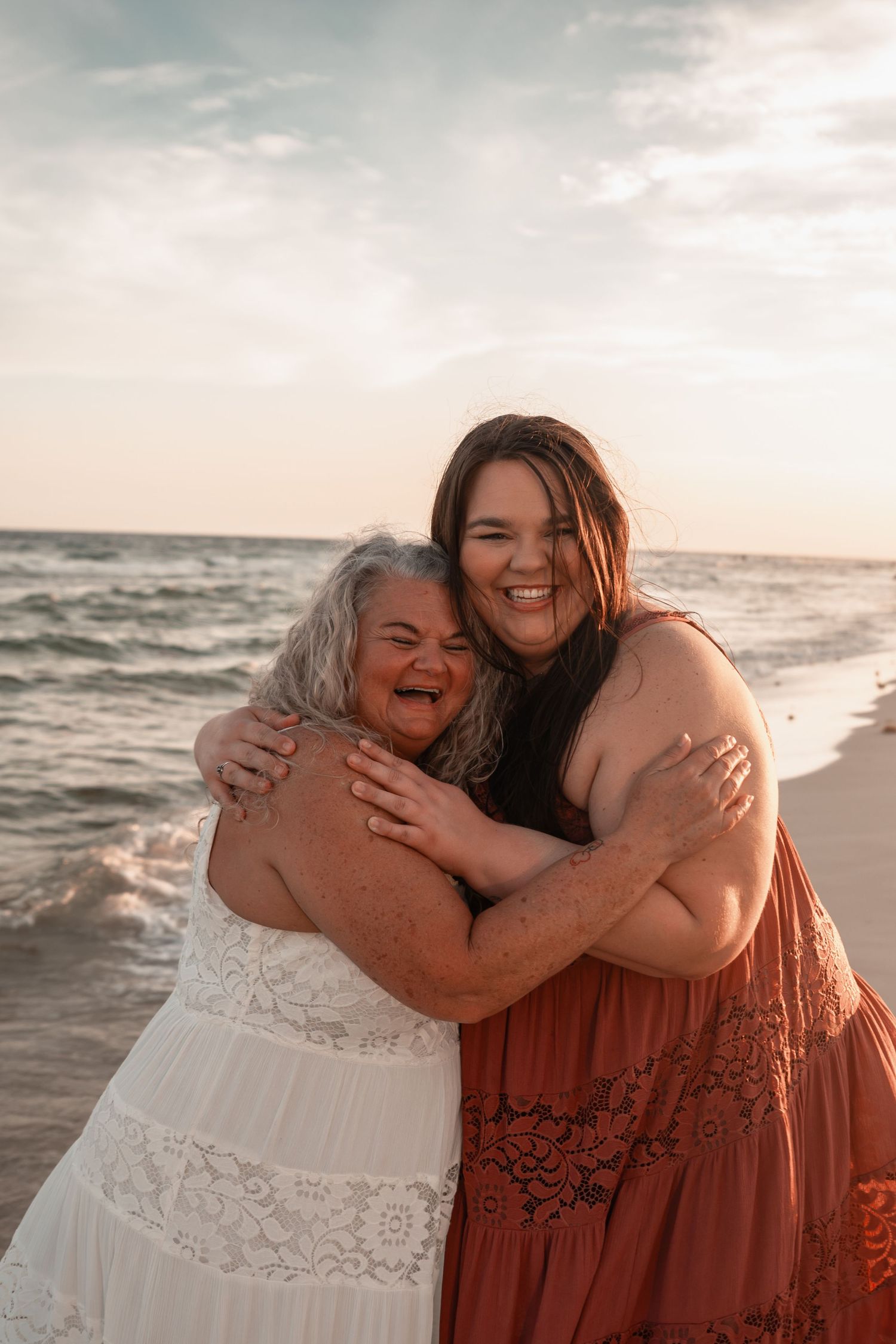 Two women hugging and laughing on a beach at sunset, one in white lace dress, other in rust boho dress.