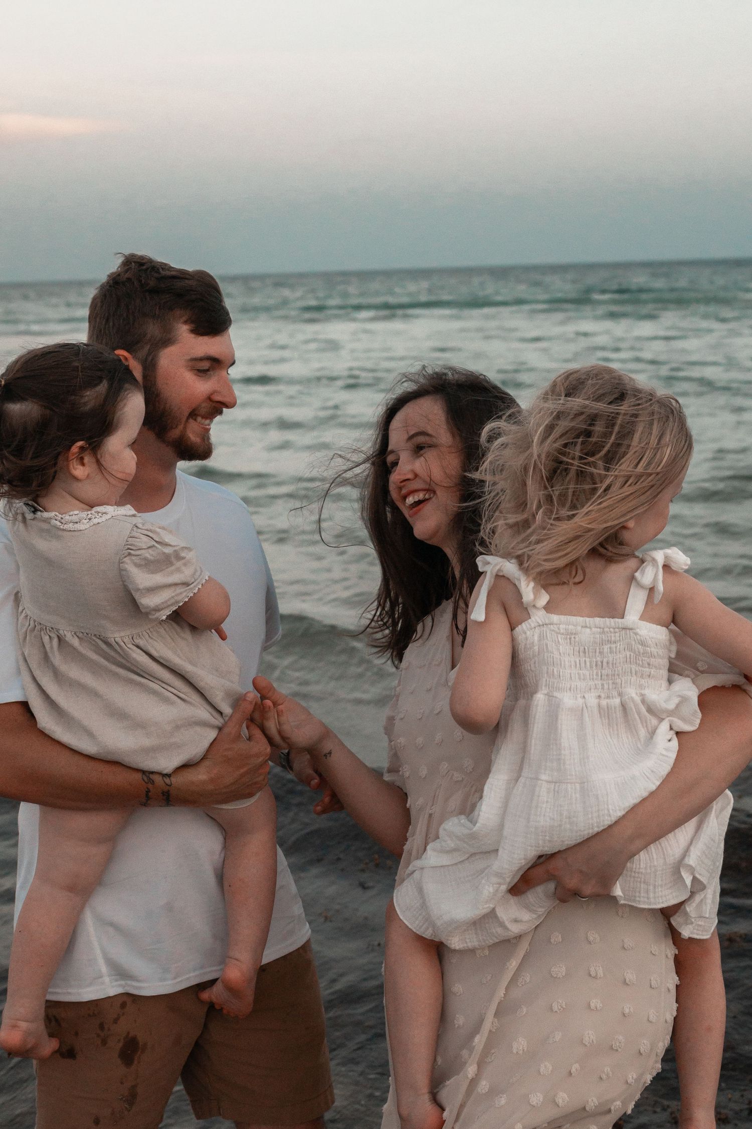 A happy young family with two toddler girls enjoys a sunset beach walk, laughing and holding hands by the ocean.