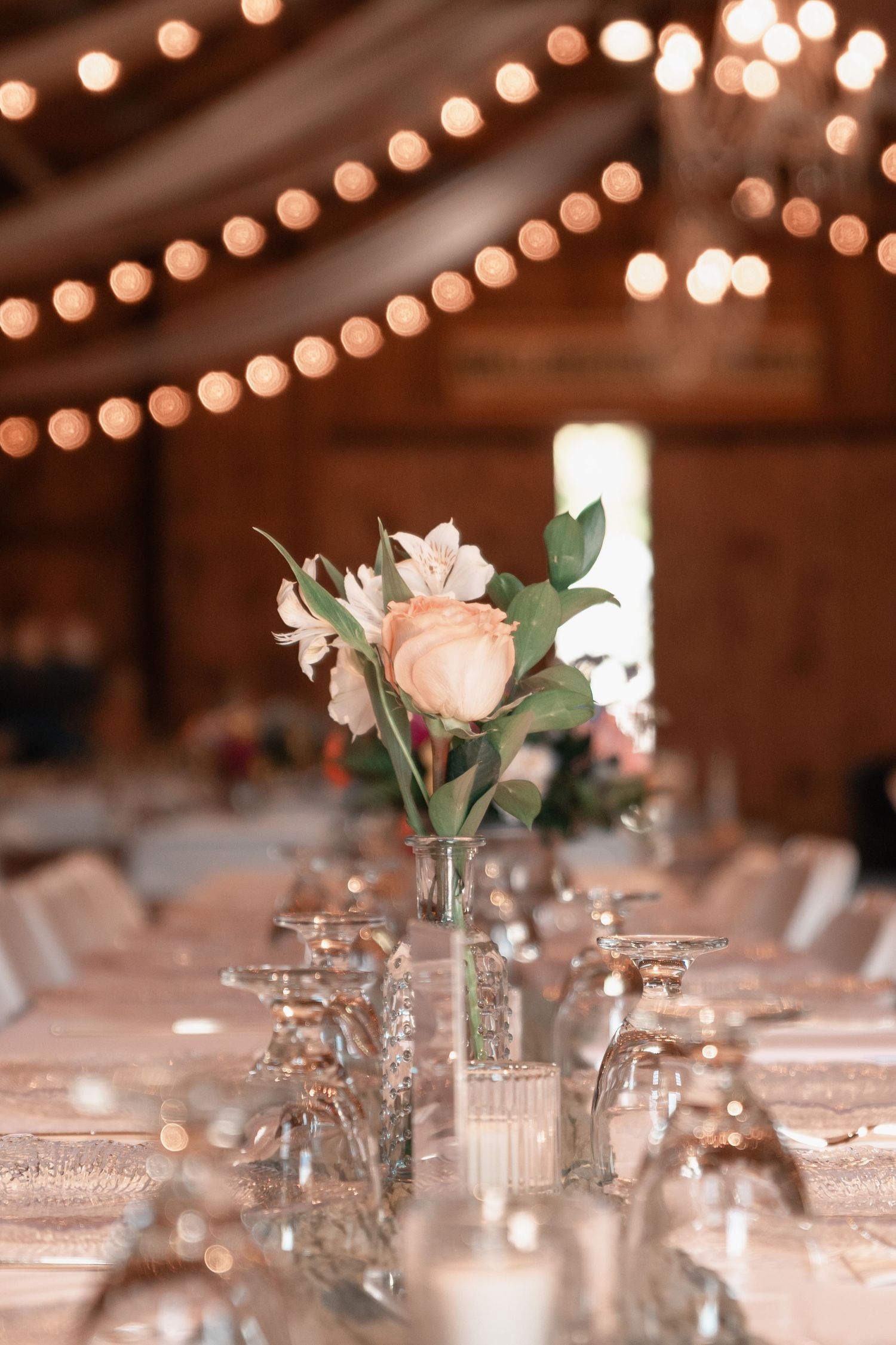 Elegant wedding reception table with peach rose centerpiece in glass vase under warm string lights.
