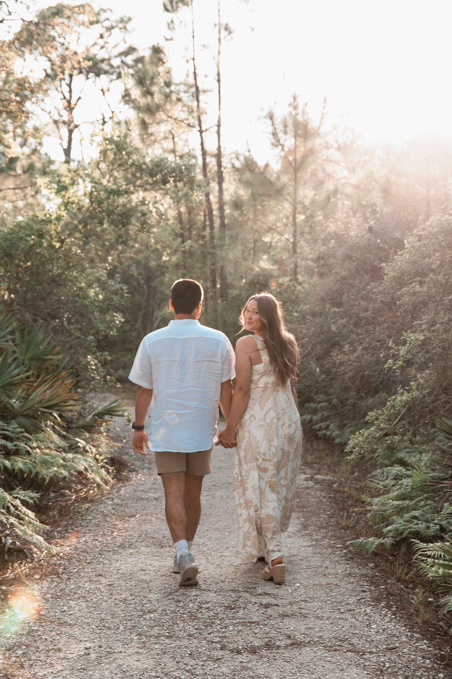 Couple strolls hand-in-hand along a sun-drenched forest path, woman in floral dress glances back smiling.