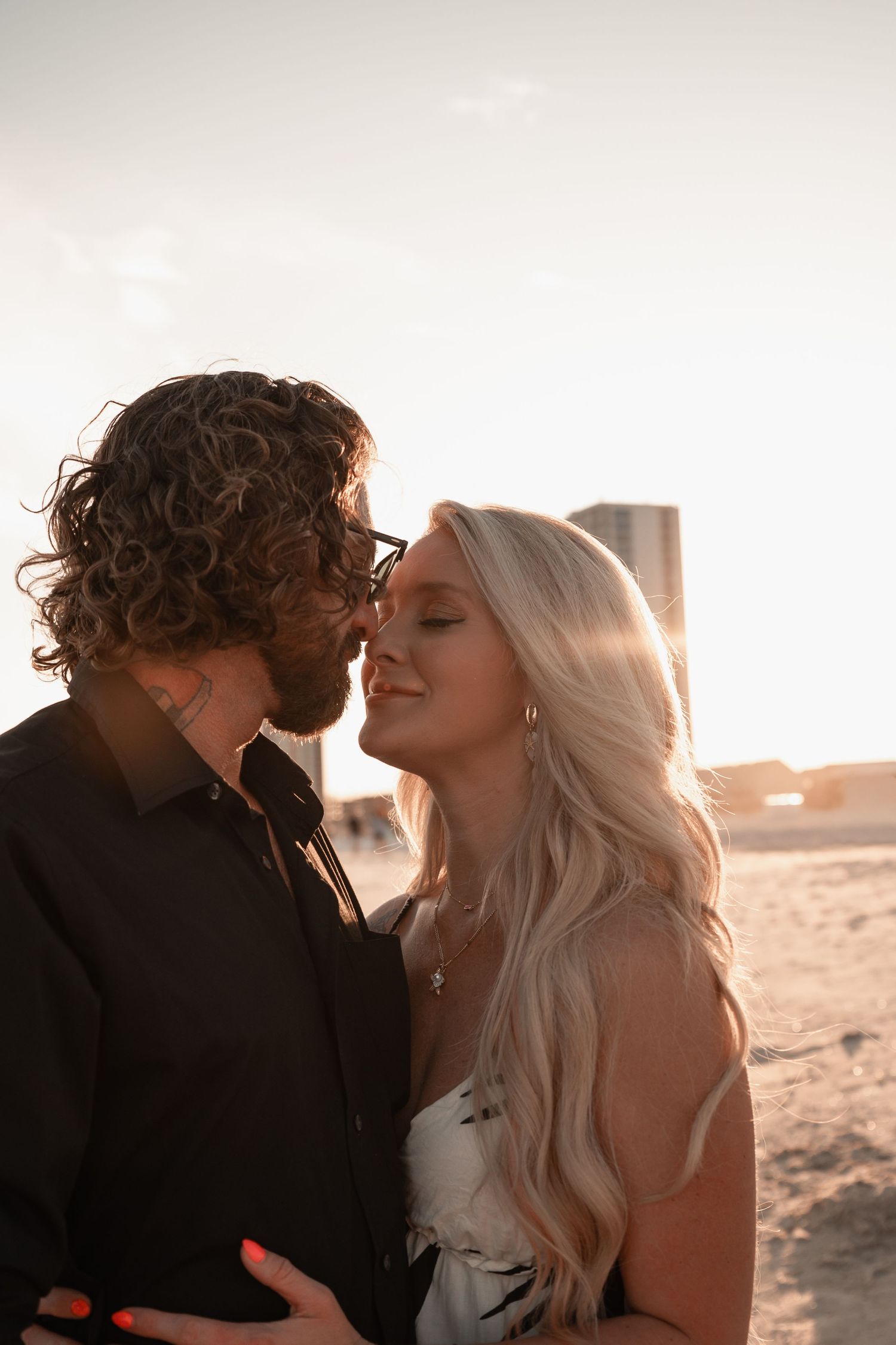 Couple nearly kissing at sunset on sandy beach, woman in white dress with long blonde hair.