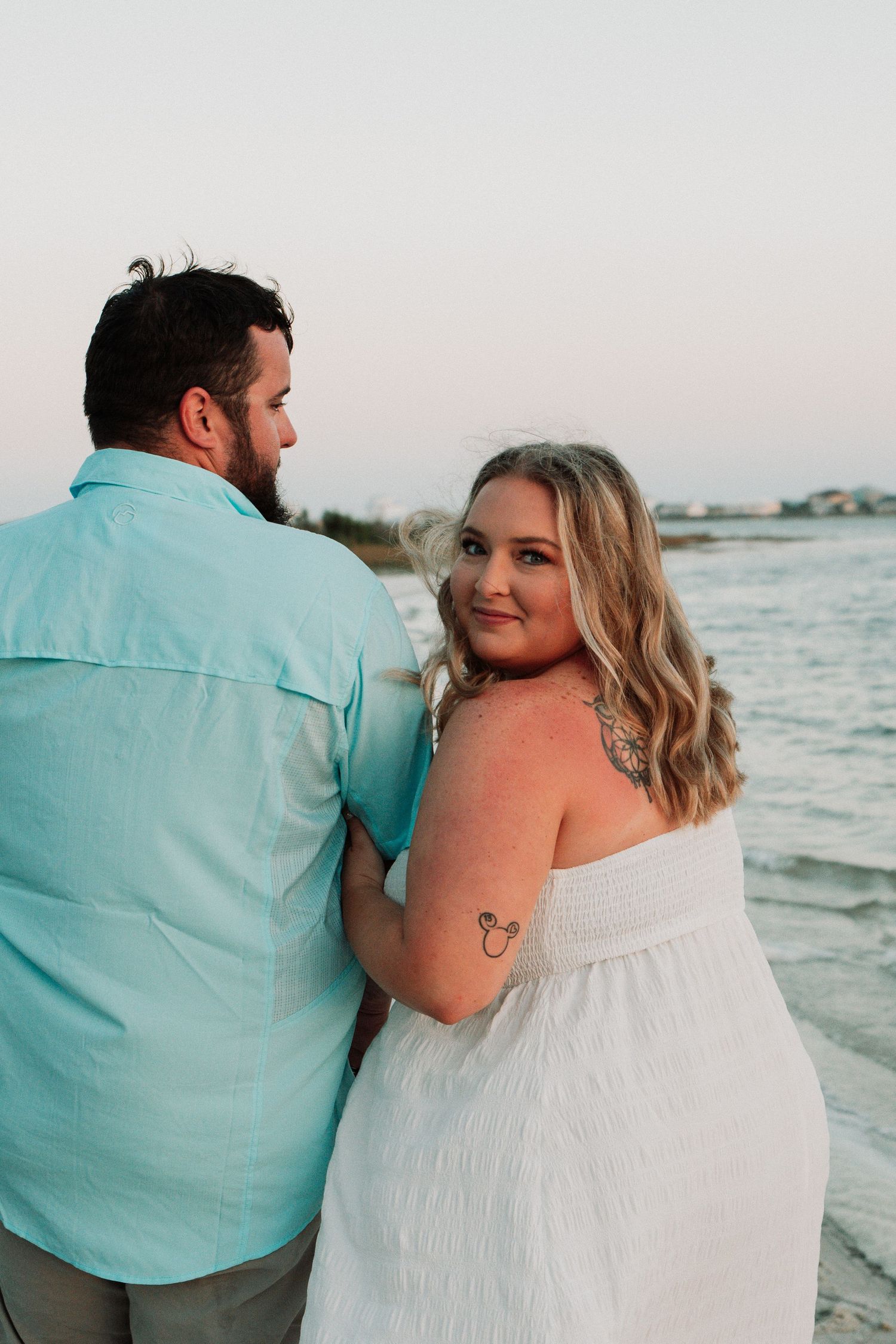 Couple posing at beach sunset, woman in white strapless dress with Mickey Mouse tattoo smiles at camera.