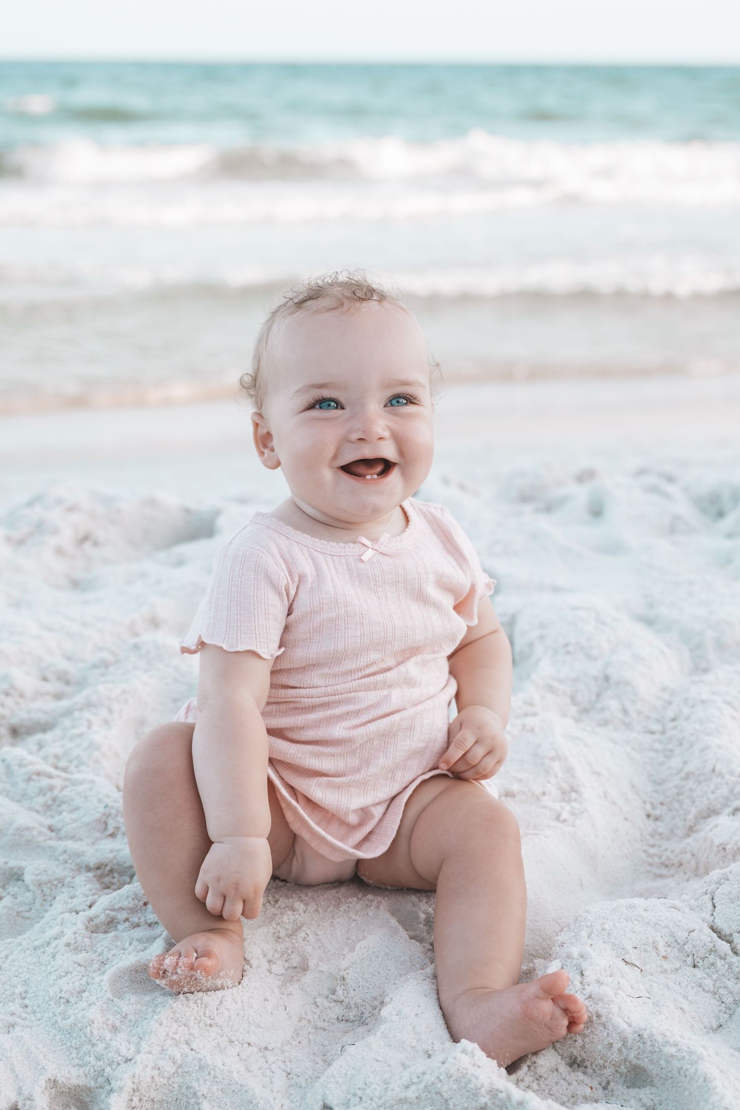 A happy baby with blue eyes sits smiling on white sand beach wearing a pink onesie with ocean waves behind.