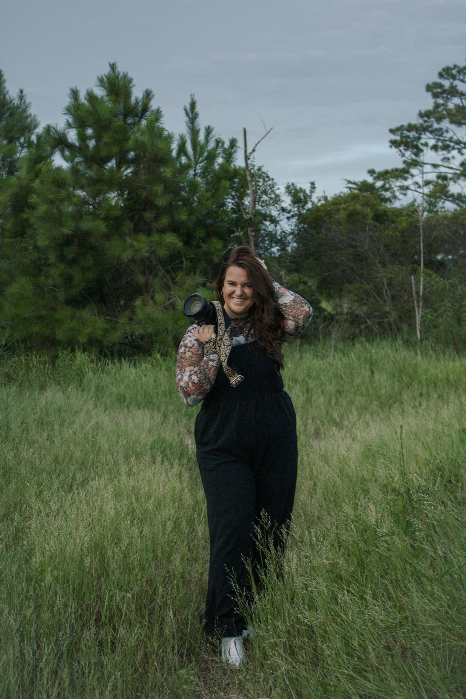 Woman in black overalls holding a camera smiling in a lush green field with trees in the background.