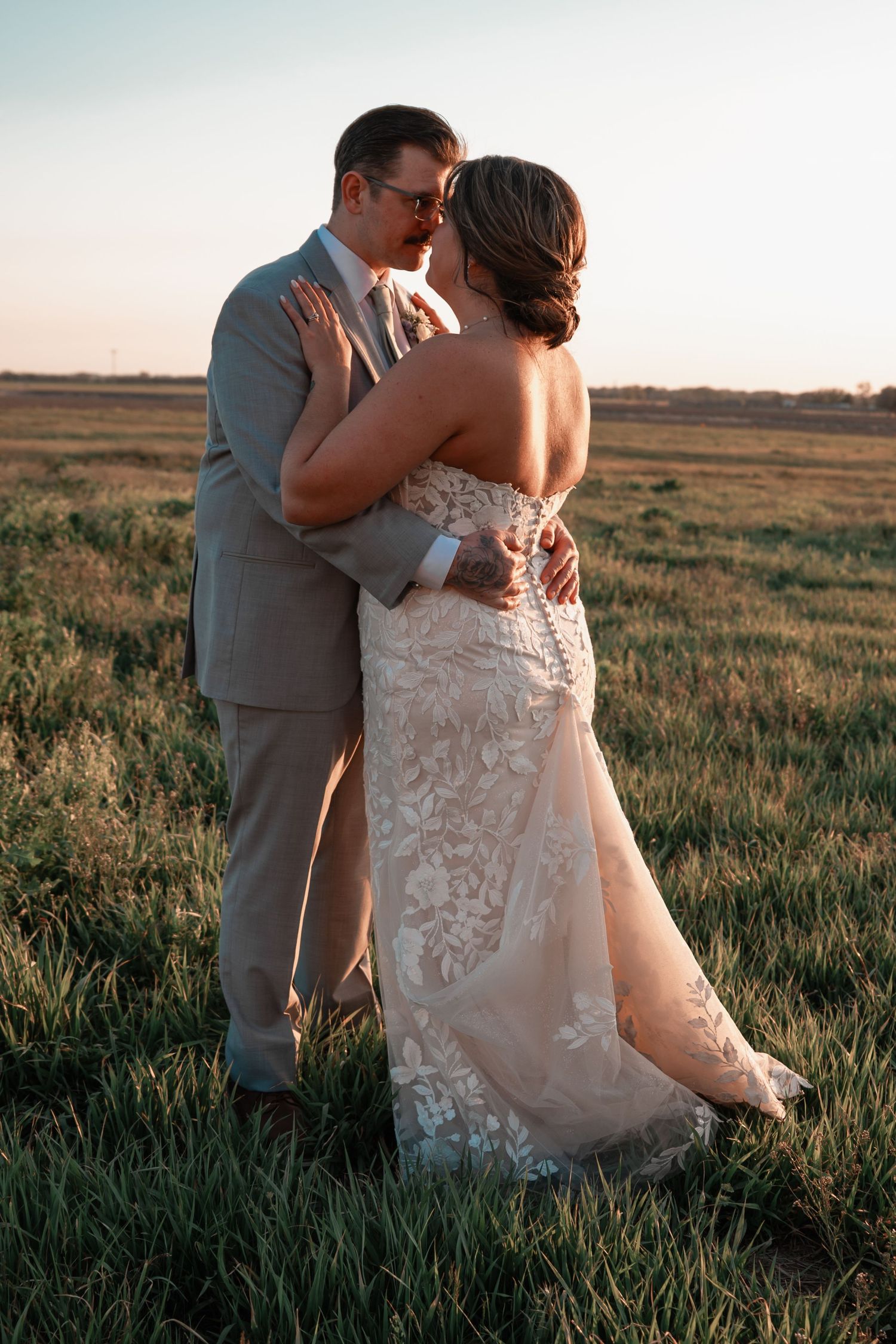 Bride and groom share romantic almost-kiss embrace in lush green field during golden hour sunset.