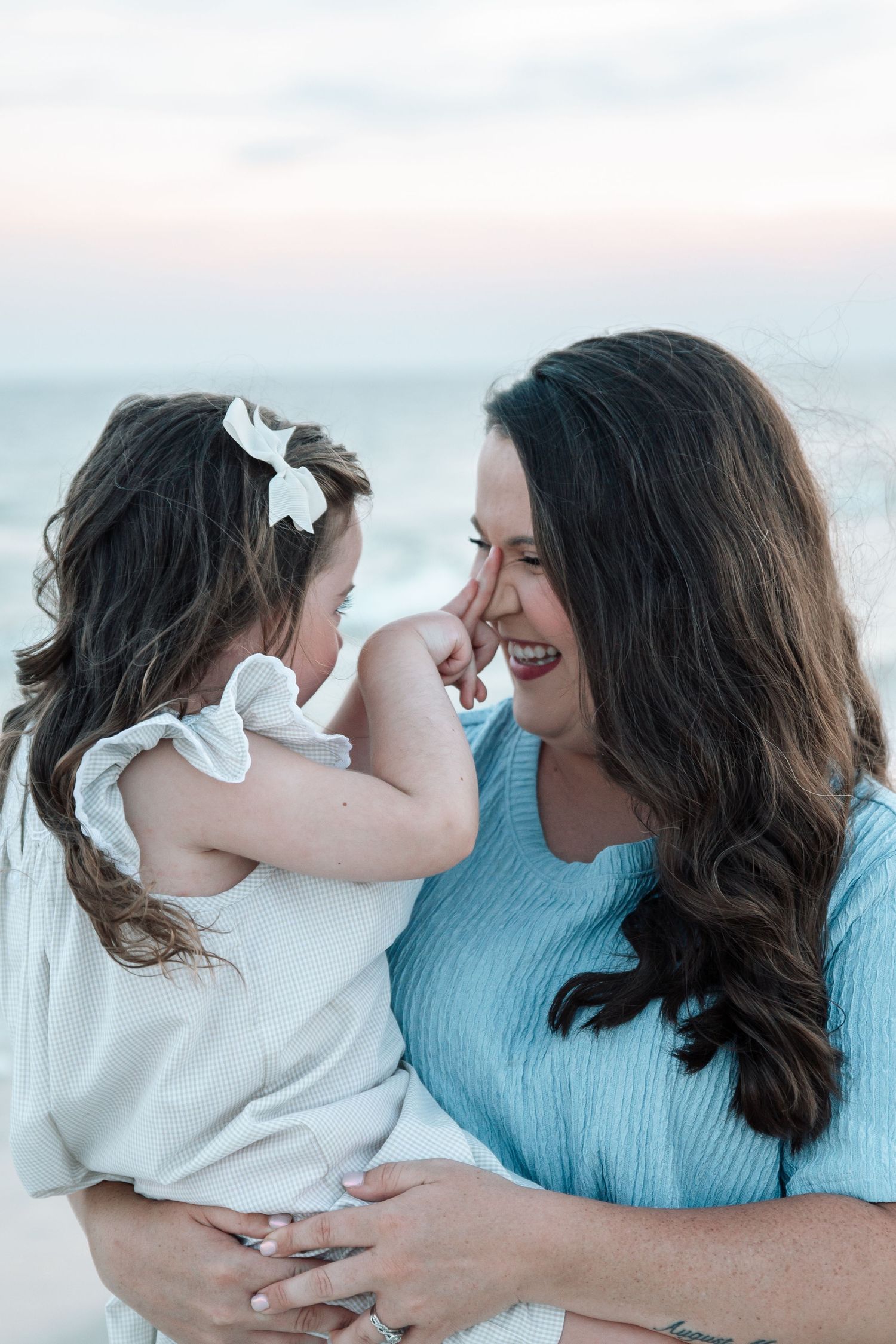 A mother holds her young daughter at the beach, both smiling and sharing a tender moment at sunset.