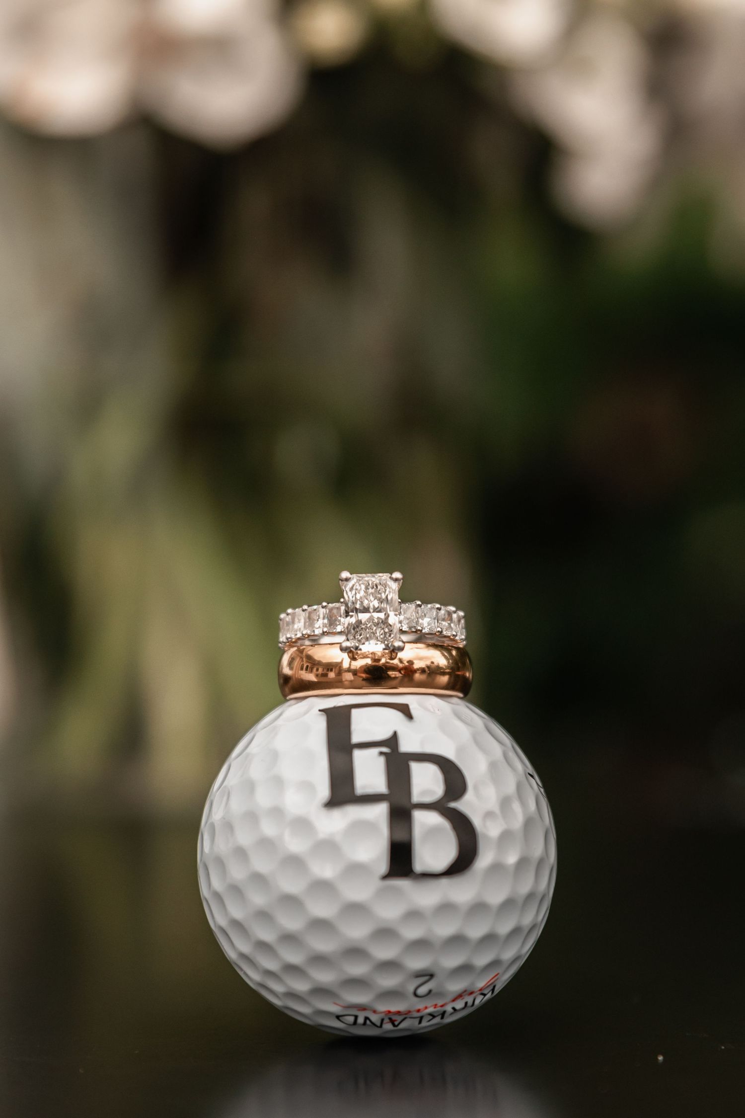 Diamond engagement ring and wedding bands displayed on a monogrammed EB golf ball with blurred greenery background.
