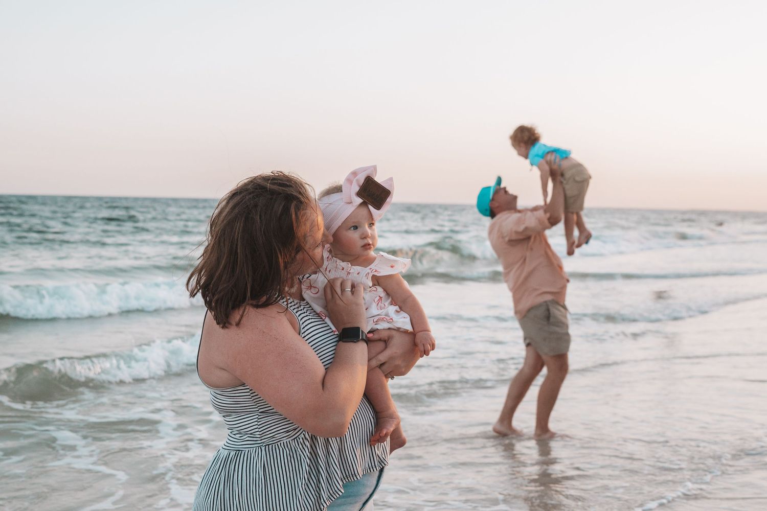 Family enjoying a beach sunset, mother holding baby girl while father lifts toddler by the ocean waves.