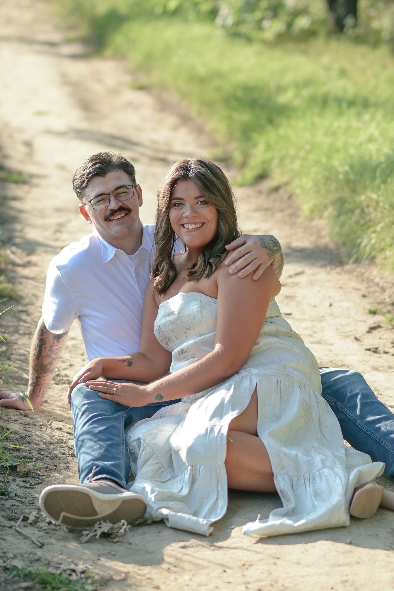 Happy couple sitting on a sunny dirt path, woman in white dress with man in white shirt and jeans.