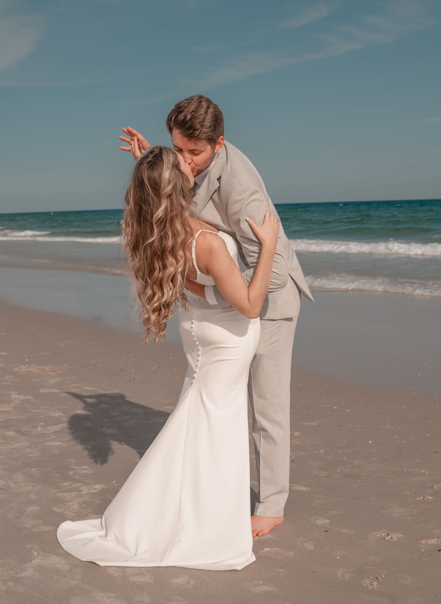Bride and groom sharing a kiss on a sunny sandy beach with ocean waves in the background.
