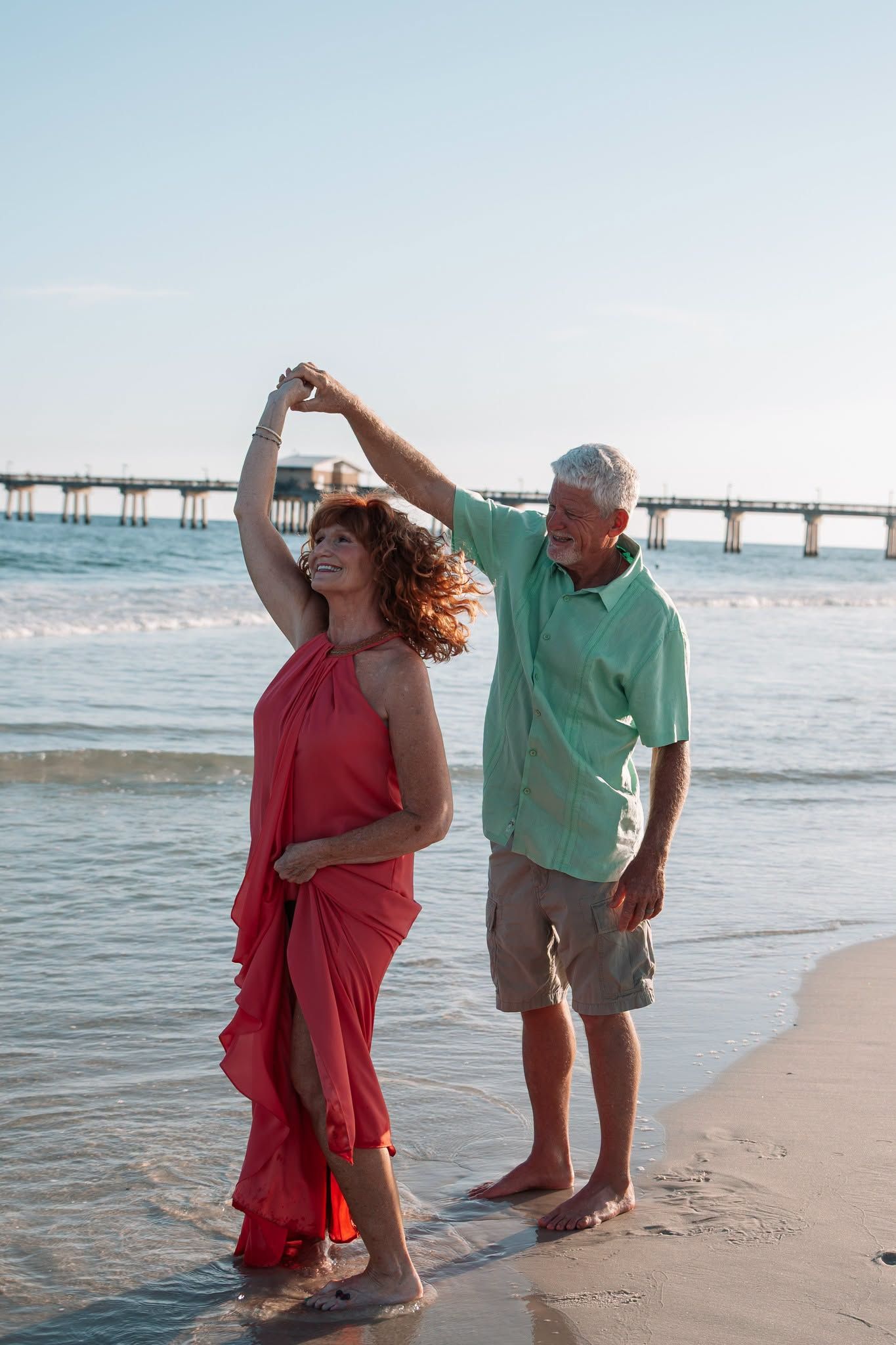 A senior couple dances barefoot on a beach at sunset, the woman in a red dress being twirled by her partner near the ocean.