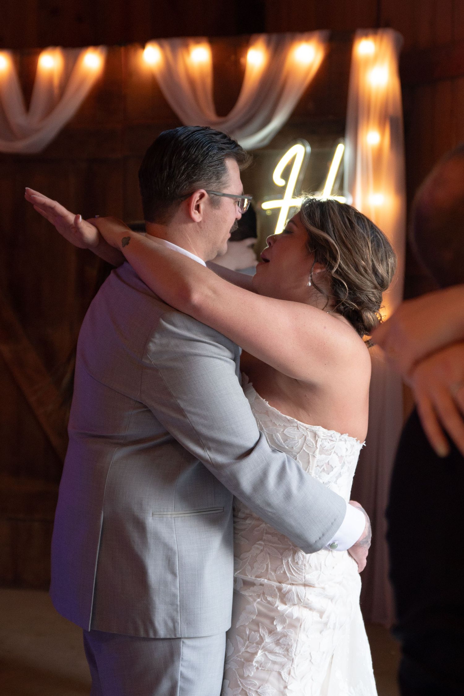 Bride and groom share intimate first dance in front of glowing neon sign at barn wedding.